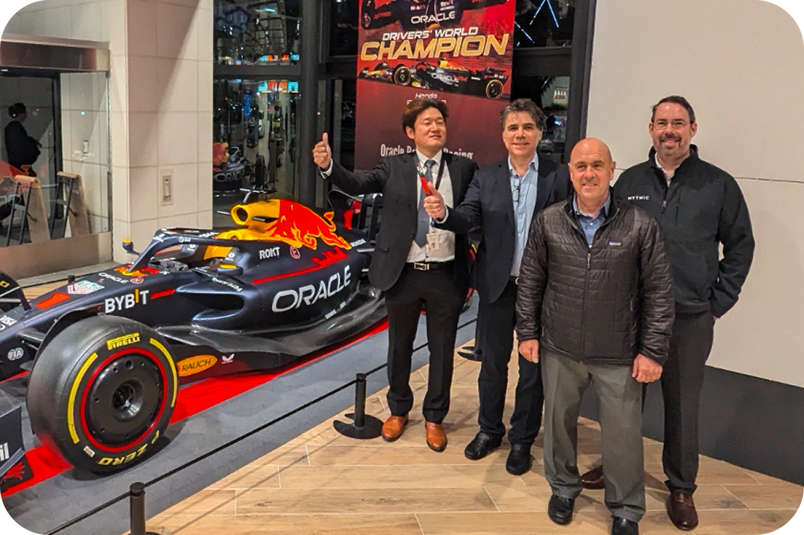 Four men standing inside near a race car with a Red Bull logo, at an event celebrating a Formula 1 driver's championship, with a banner in the background.