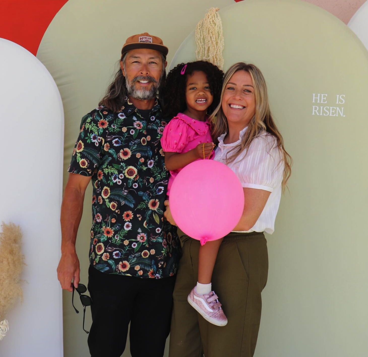 A man, woman, and young girl smiling and posing together at a celebration. The girl is holding a pink balloon and wearing a pink dress, while the man and woman are dressed casually. They are standing in front of a pastel-colored backdrop with the partial phrase 'HE IS RISEN.'