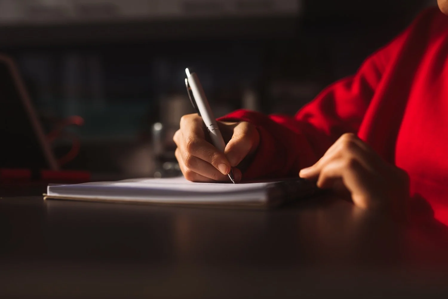 Person writing in a notebook with a pen, wearing a red shirt, in a dimly lit room.