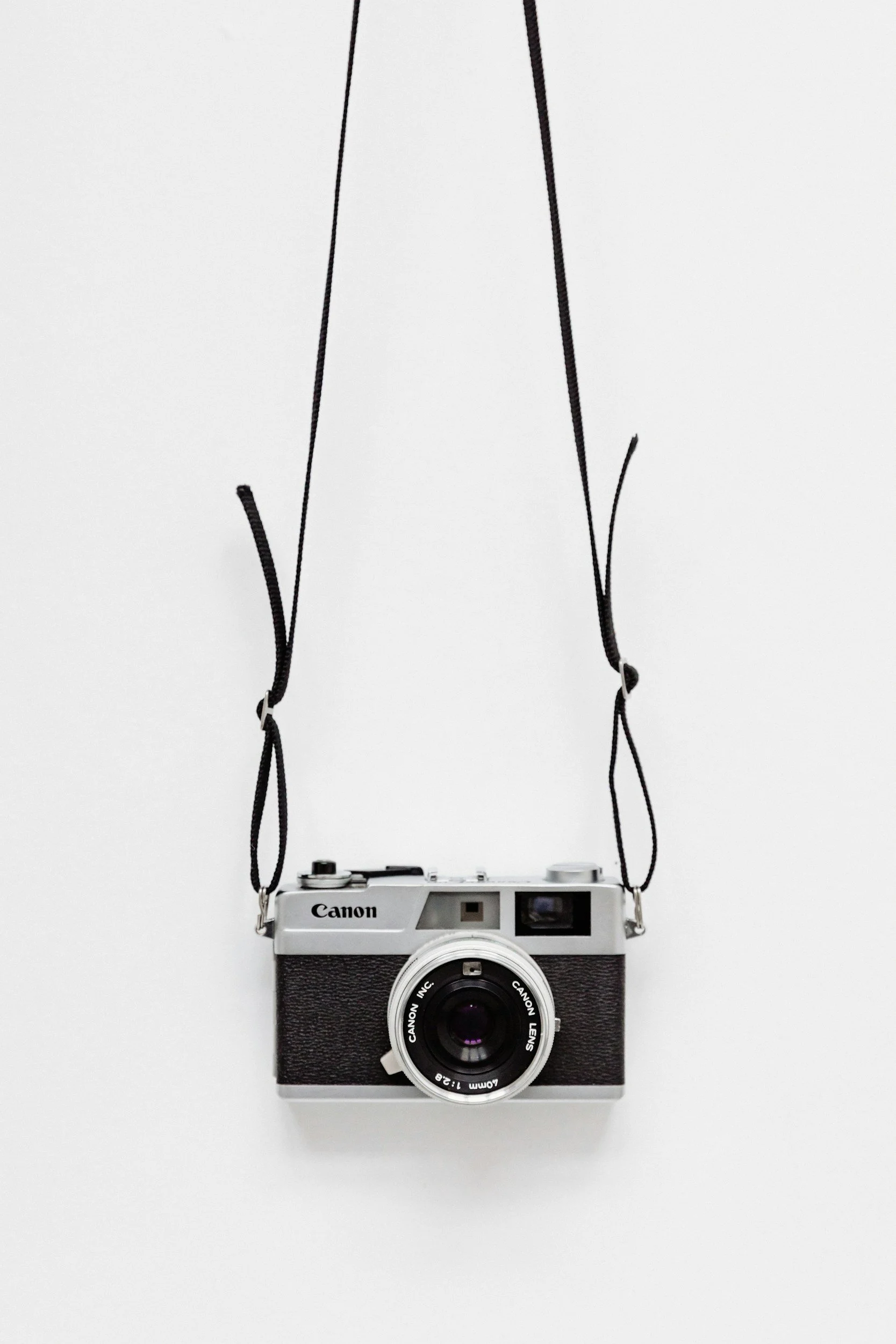 Silver and black Canon vintage camera with a black strap hanging against a white background.