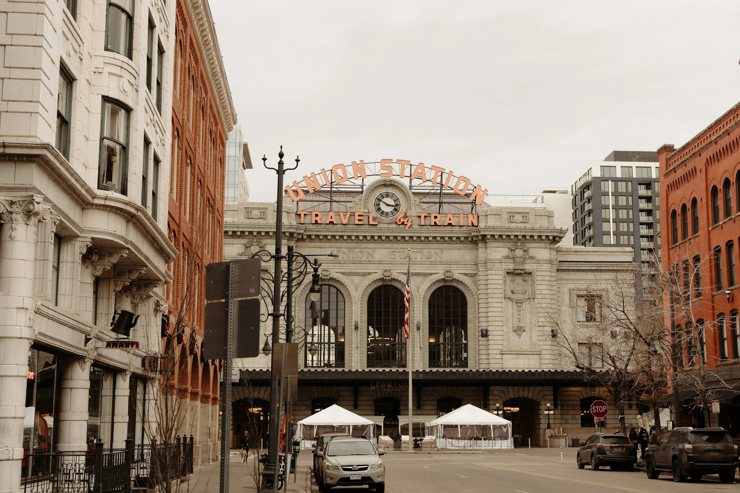 The image shows Union Station with the sign reading 'Travel by Train' on top of the building. There are several cars parked in front, and the street is lined with historic buildings. The sky is overcast.