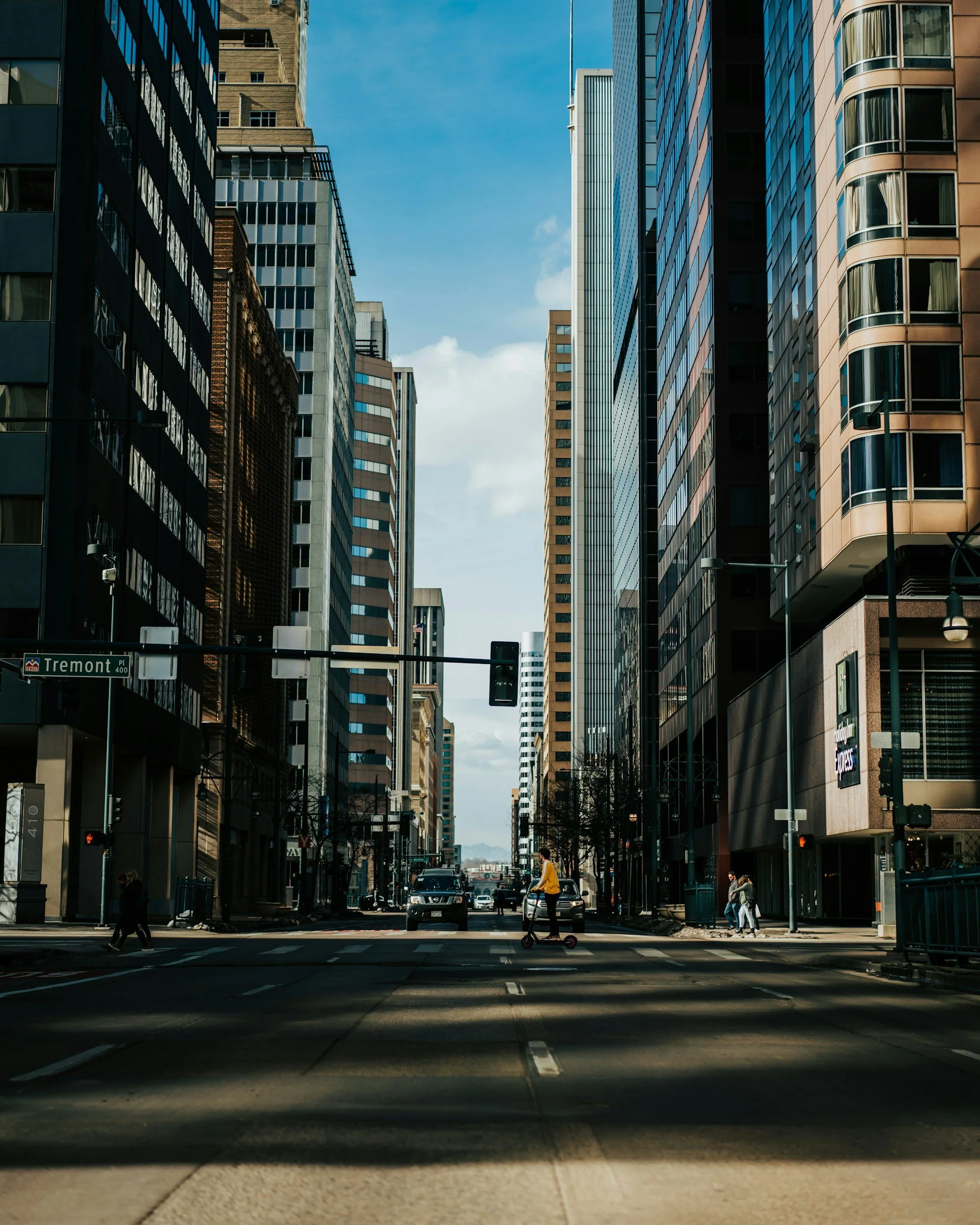 A city street scene with tall office buildings on both sides and a clear blue sky overhead. Pedestrians cross the street, and cars drive along the road. A man in a yellow jacket and a person on a scooter are visible in the foreground.