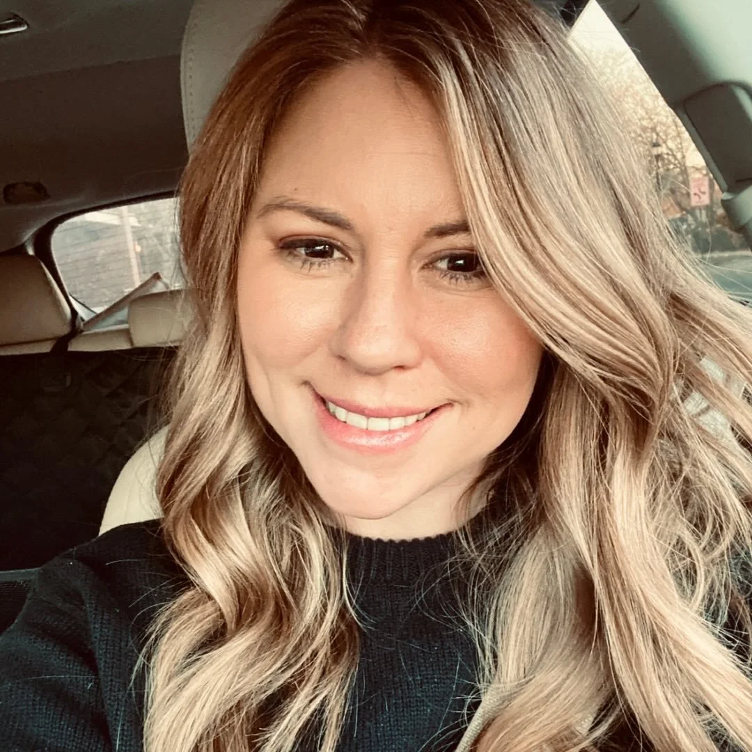 A woman with long wavy blonde hair smiling inside a car.