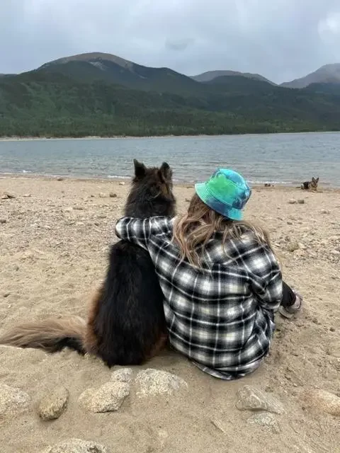 A girl and a dog sitting on the sandy beach, facing a lake with mountains in the background.