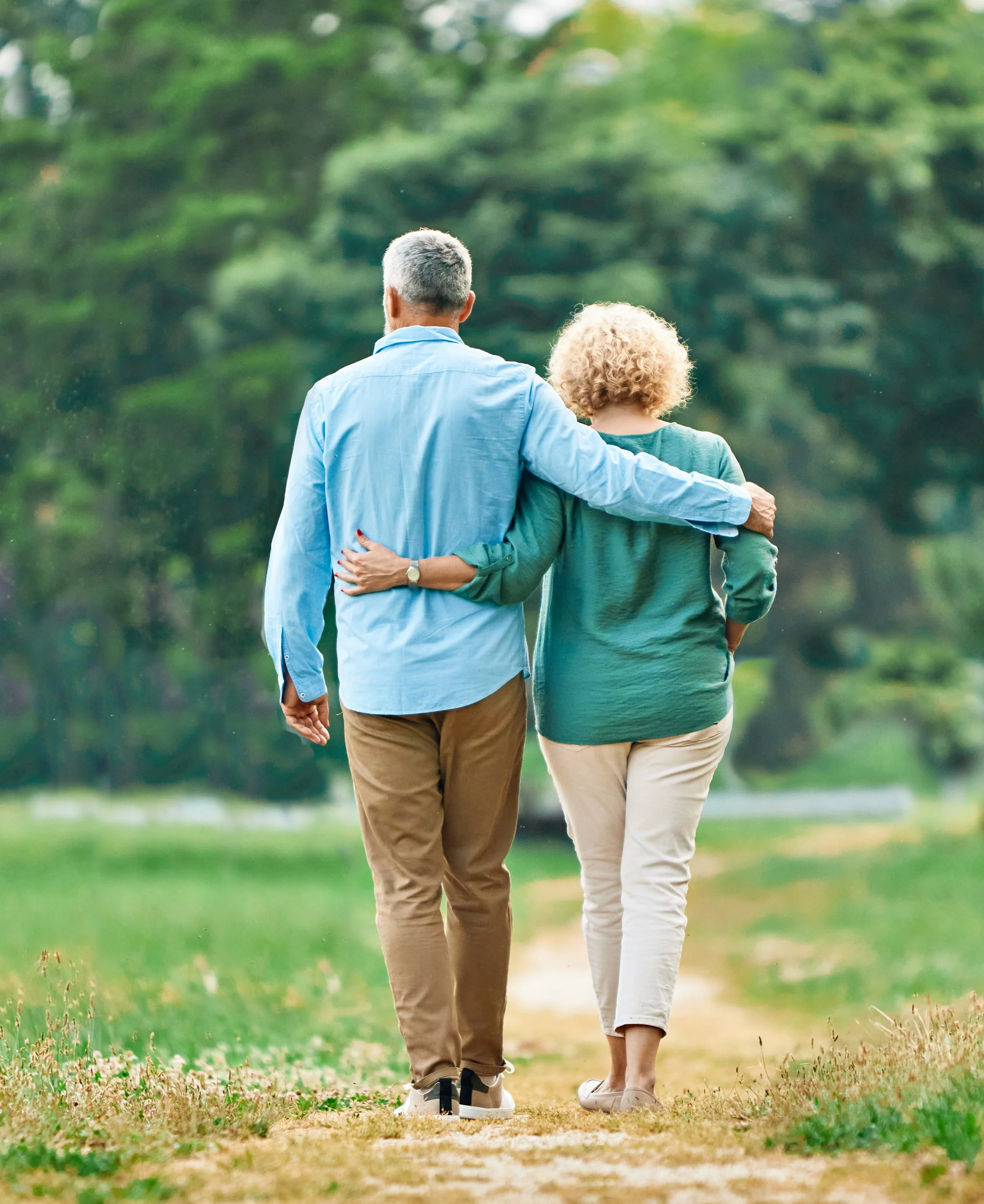 Two people walking together in a park, with one arm around each other's shoulders, viewed from behind. They are walking along a path amidst green grass and trees.