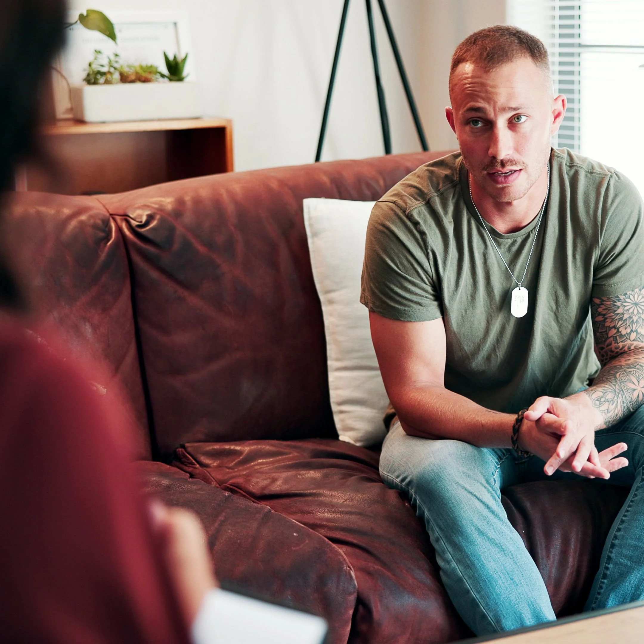 A young man with short hair, tattoos on his arm, and a dog tag necklace sitting on a leather couch in a well-lit room with blinds, plants, and a lamp in the background, looking to his right.
