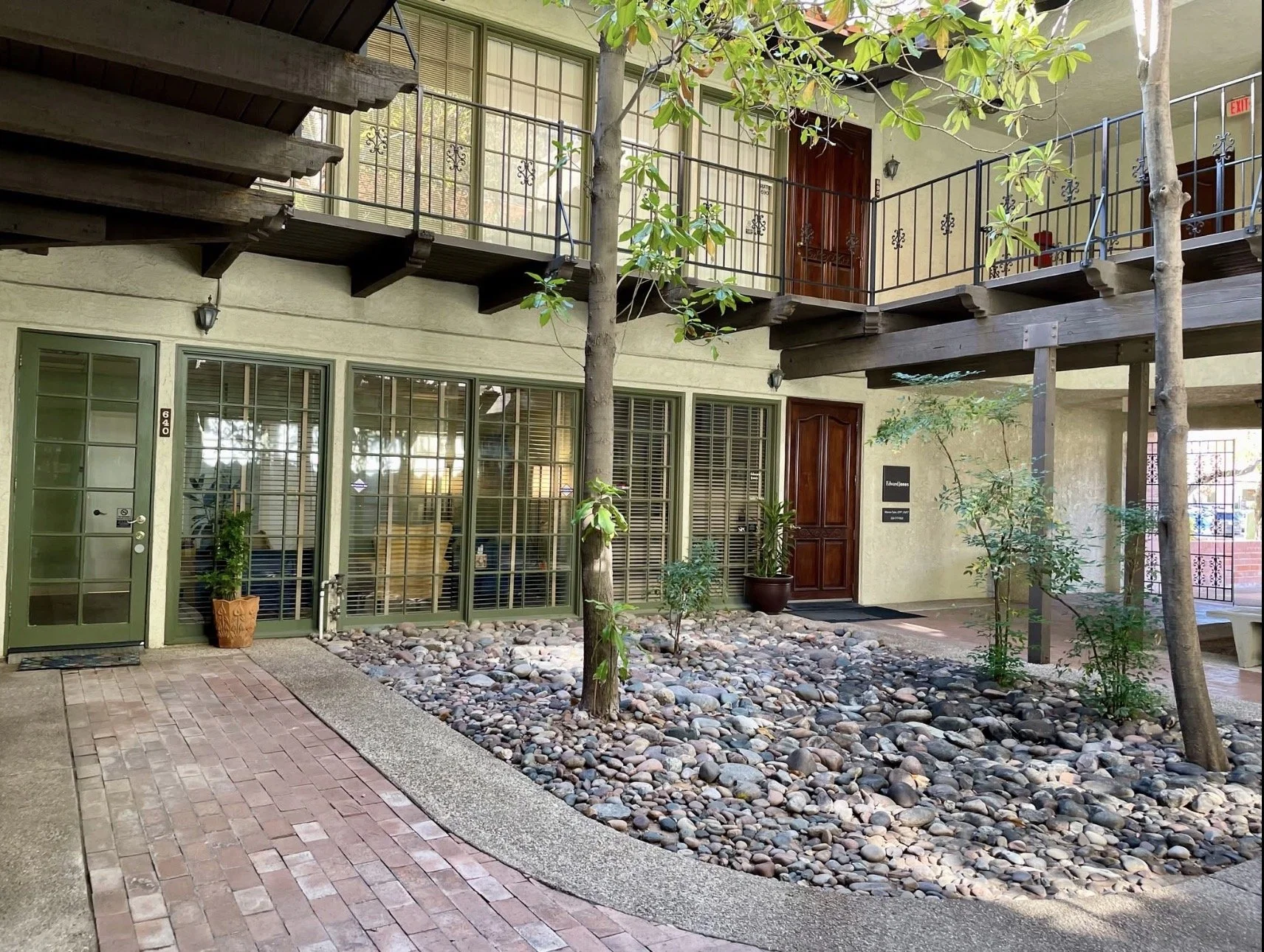 Residential apartment building with a ground floor entrance and an upper balcony. The ground floor has green-framed windows and doors, including a glass door and wooden door. In front, there is a small landscaped area with rocks and young trees. The balcony has a black metal railing. There are potted plants near the entrance.