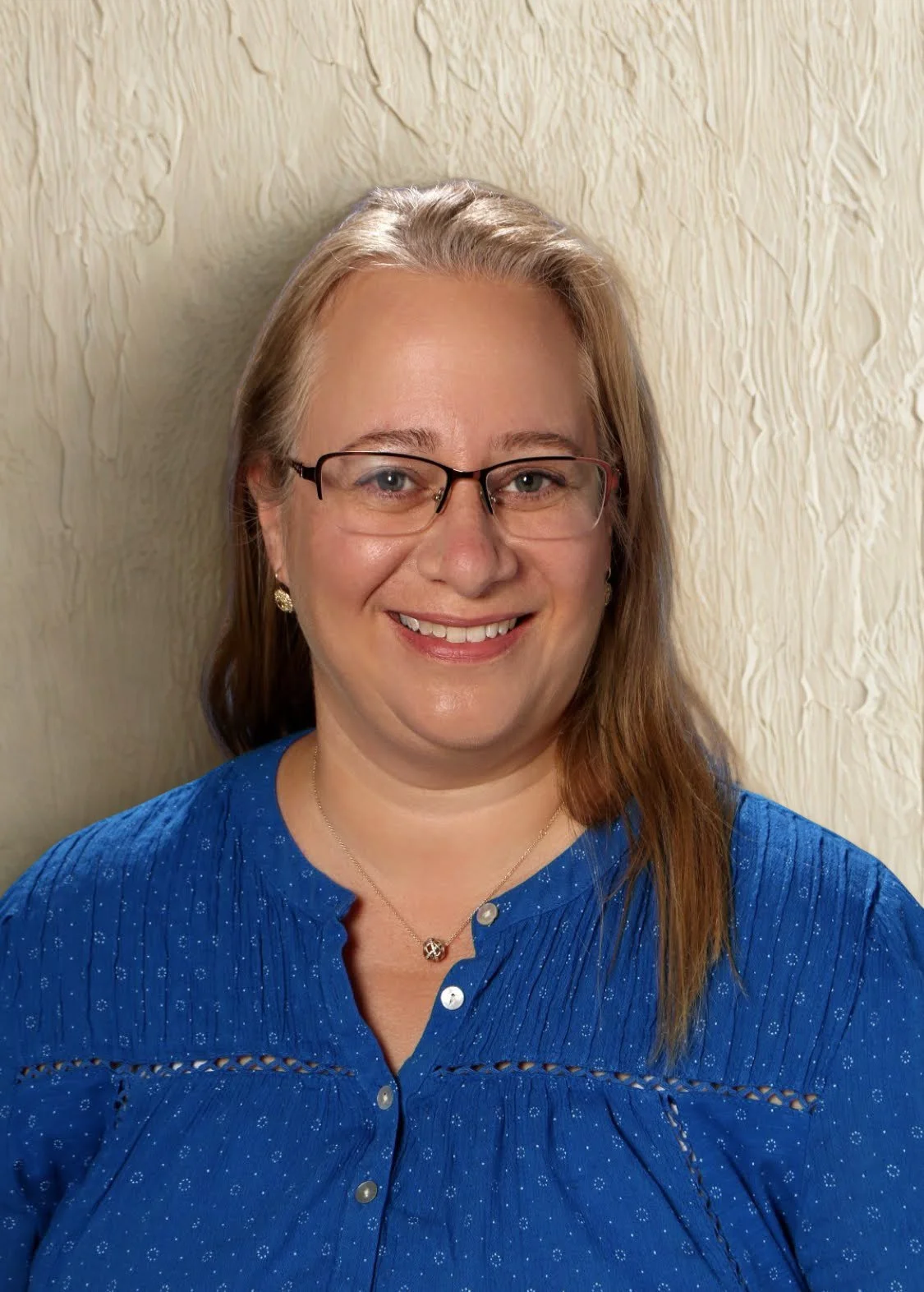 A woman with glasses and shoulder-length brown hair smiling at the camera, wearing a blue blouse and a necklace with a small pendant, in front of a light textured wall.