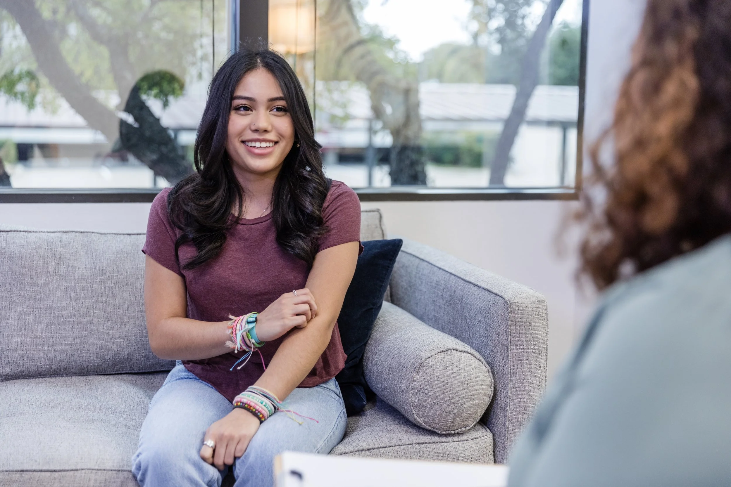 A young woman with long dark hair, smiling, sitting on a beige couch, wearing a maroon t-shirt and light blue jeans, engaging in conversation with another person who has curly hair, in a room with large windows showing trees outside.