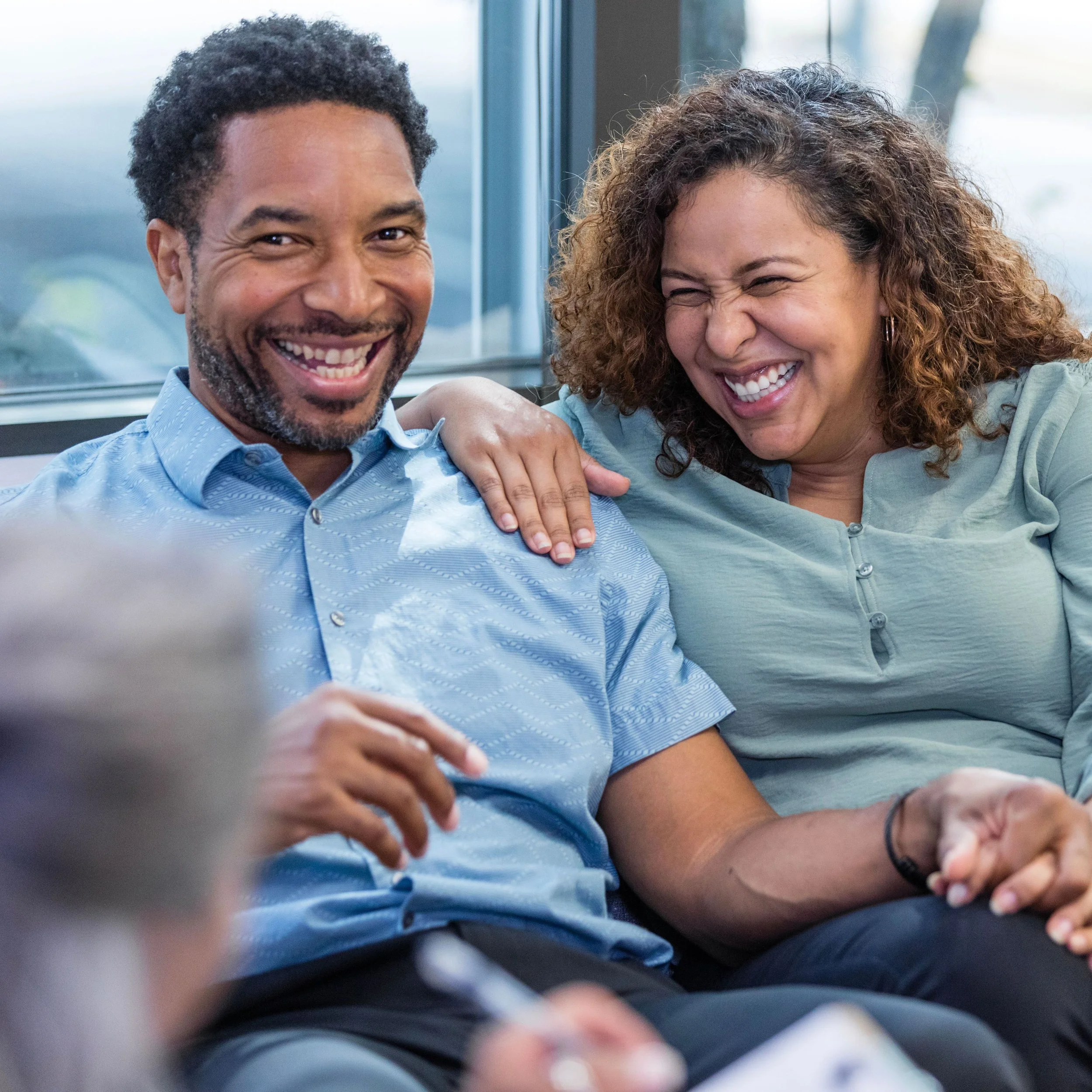 A close-up of a smiling man and woman, sitting close together, with the woman touching the man's shoulder in a bright room with large windows in the background.