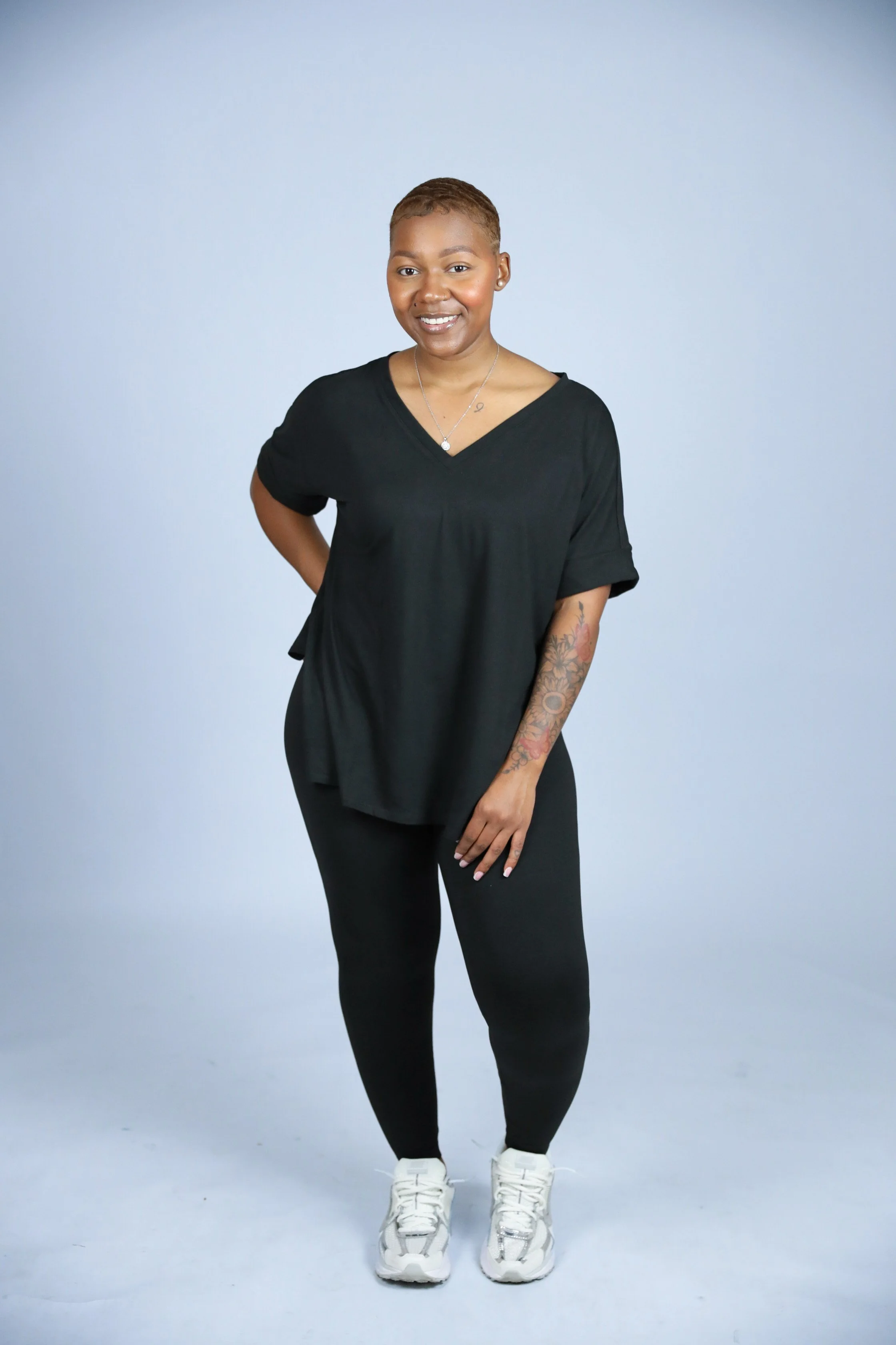 A smiling woman with short hair, wearing a black V-neck top, black pants, white sneakers, jewelry, and showing a colorful tattoo on her left arm, standing against a plain light background.