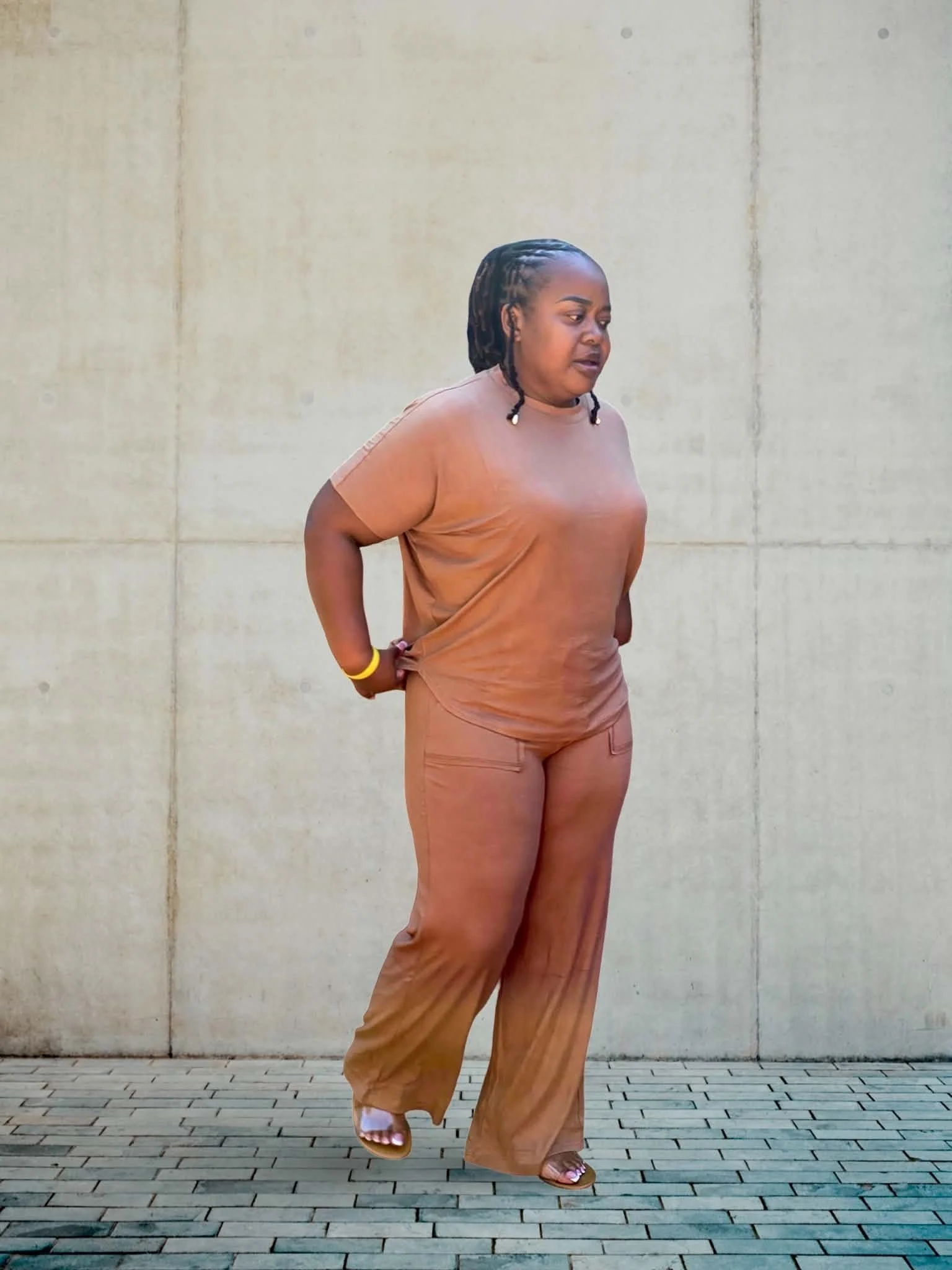 A woman with dark hair styled in braids, wearing a tan T-shirt and matching loose pants, standing against a beige concrete wall, looking to the side with a neutral expression.
