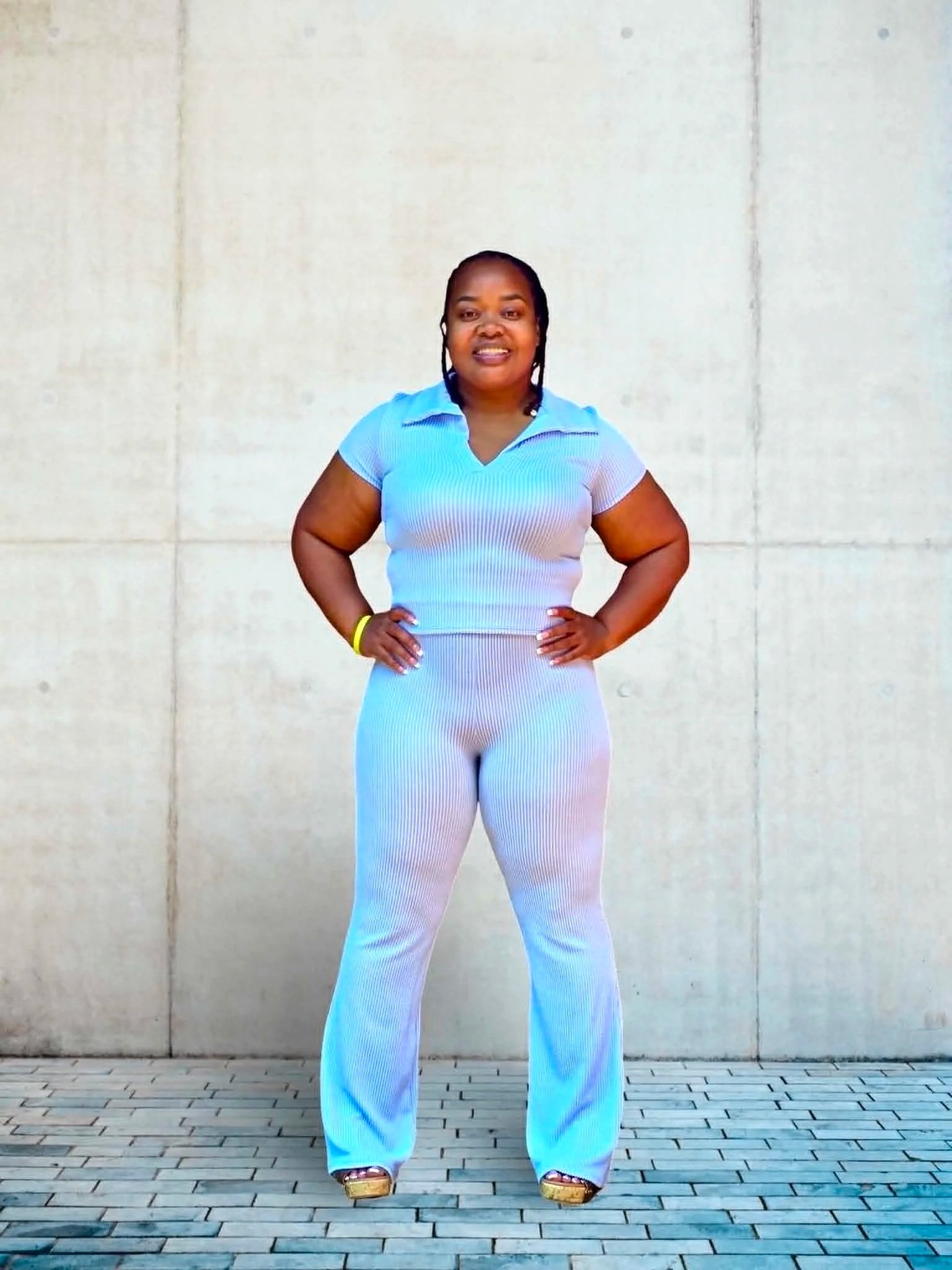 A woman with dark skin, wearing a blue and white striped shirt and light-colored pants, stands with her hands on her hips in front of a plain, light-colored wall. She has dark braided hair and is wearing shoes with thick heels.