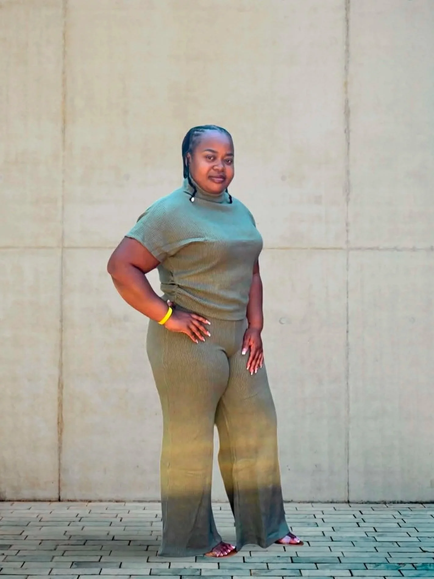 A woman in a tan outfit with wide-legged pants and a short-sleeve top stands outdoors against a beige wall, with her left hand on her hip and her right arm relaxed at her side.