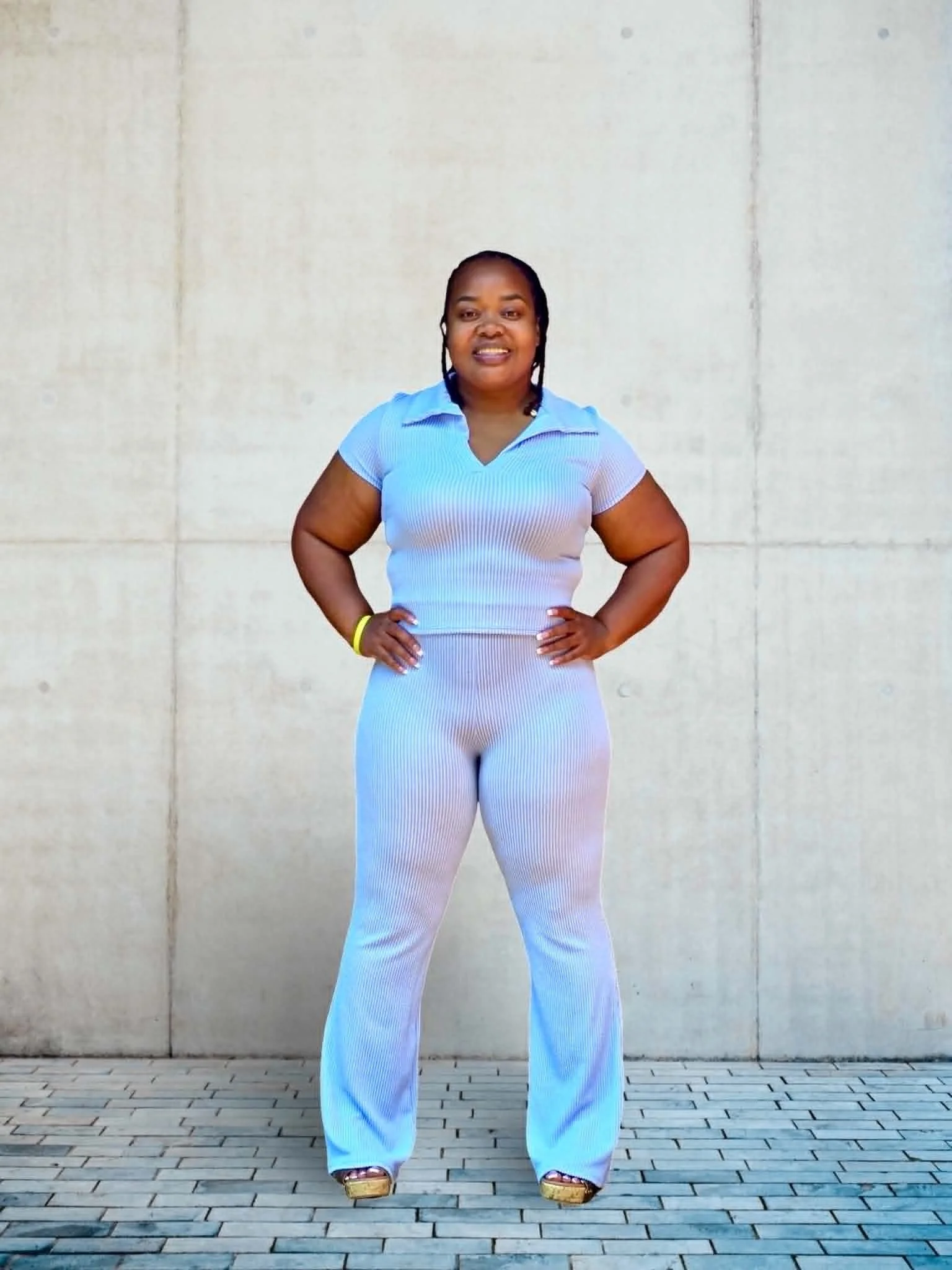 A woman with dark hair in braid, standing outside in front of a plain concrete wall. She is wearing a light blue fitted polo shirt and white wide-legged pants, with her hands on her hips. She has a yellow wristband on her right wrist and is wearing sandals with a platform heel. The ground is covered with light-colored brick pavers.