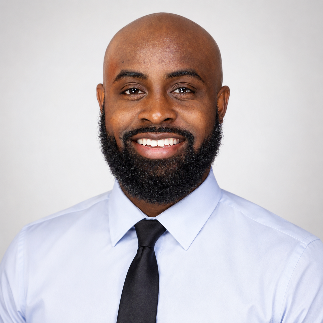 Headshot of a smiling black man with a bald head and full beard, wearing a light blue dress shirt and black tie against a neutral background.