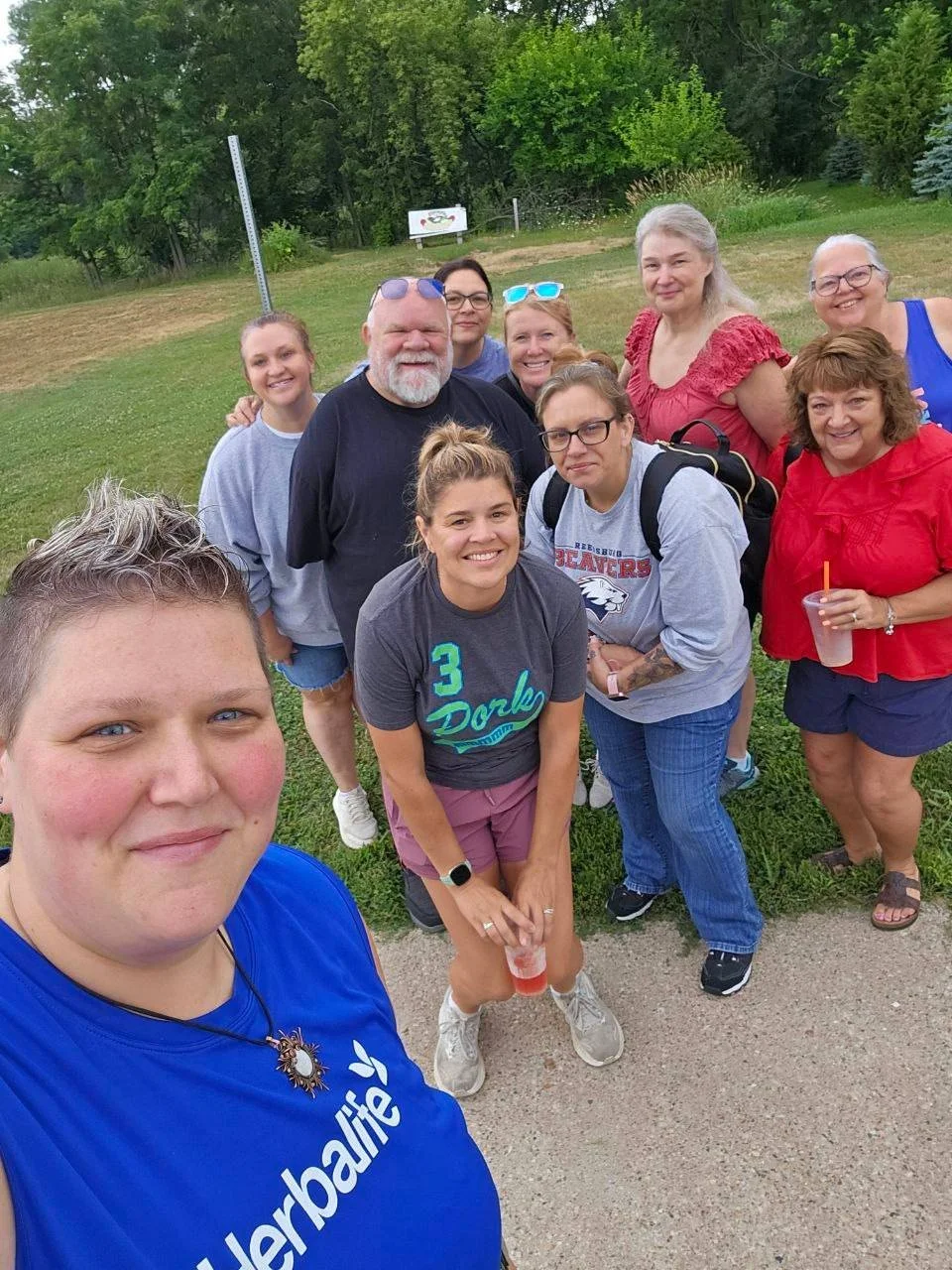 A group of people posing outdoors, smiling for a selfie. They are standing on a grassy area with trees in the background and appear to be at a casual gathering or walk.