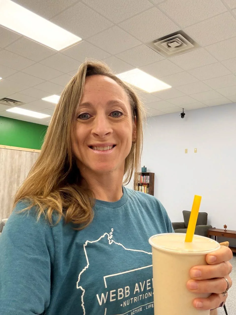 A woman with long, light brown hair smiling and holding a cup with a yellow straw in an indoor room with a drop ceiling, fluorescent lights, and light blue walls.