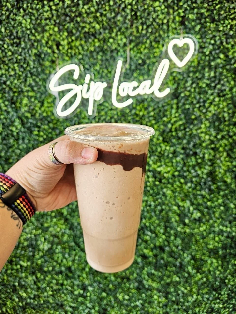 Hand holding a blended chocolate milkshake with chocolate syrup drizzled on top, in front of a green artificial hedge wall with a neon sign that says 'Sip Local' and a heart symbol.