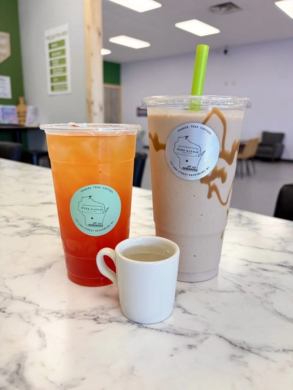 Two large beverages in plastic cups and a small ceramic cup with tea on a marble table. The left beverage is an orange-colored iced drink, and the right is a light brown iced coffee with caramel drizzle. Both cups have a blue circular logo with a map outline and text, indicating they are from Webb Avenue in Reedsburg, Wisconsin.