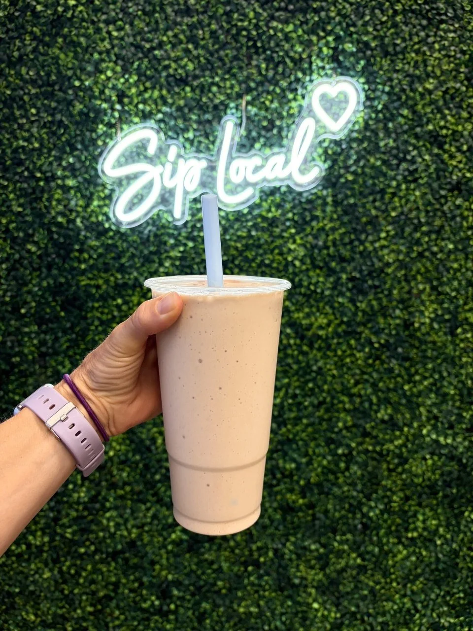 Person holding a large smoothie drink in front of a green artificial hedge wall with a neon sign that says 'Sip Local' with a heart.