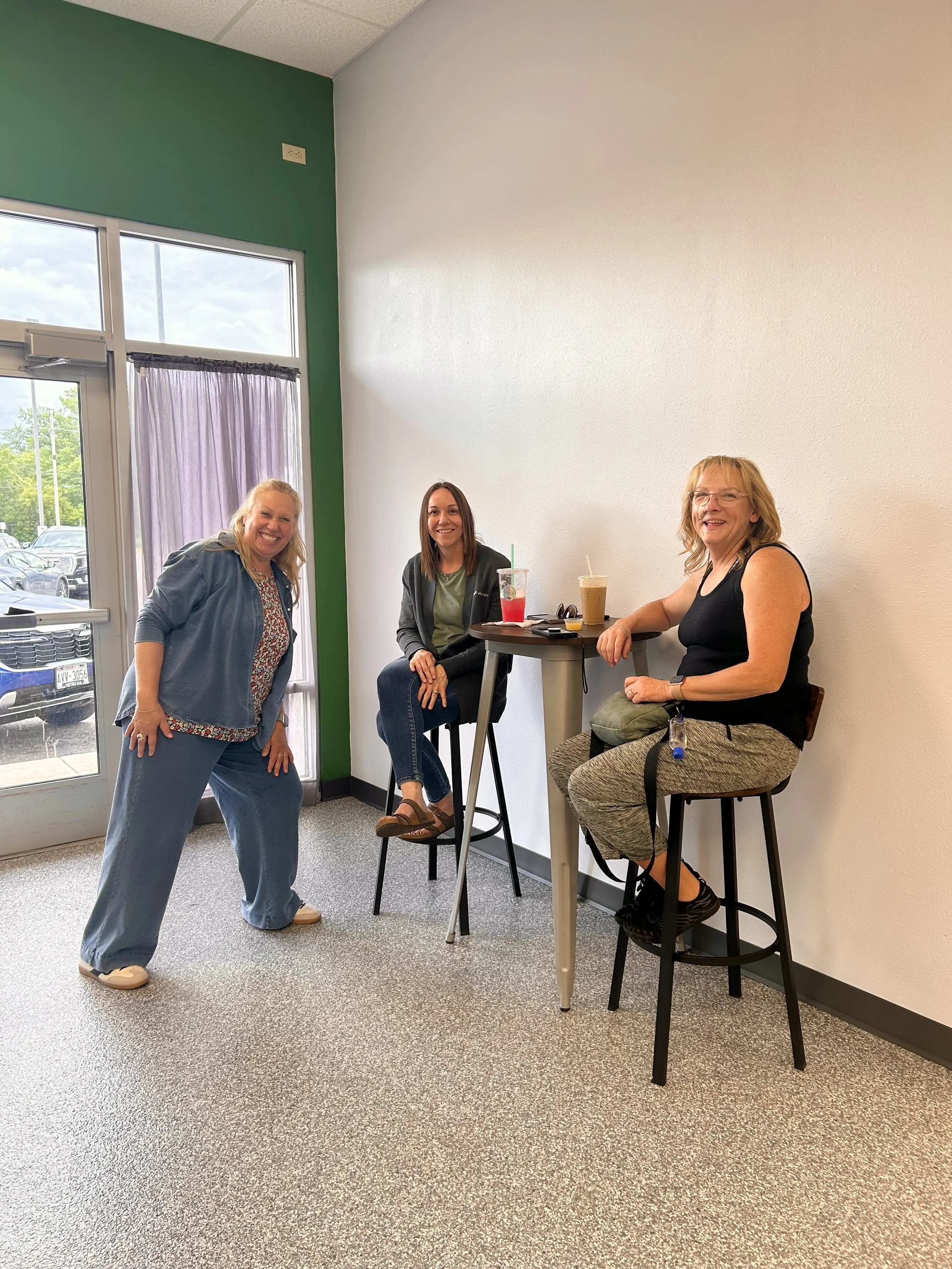 Three women in a casual setting, two sitting on high chairs at a round table with drinks, one standing next to them smiling. The room has a glass door and a white wall, with natural daylight outside.