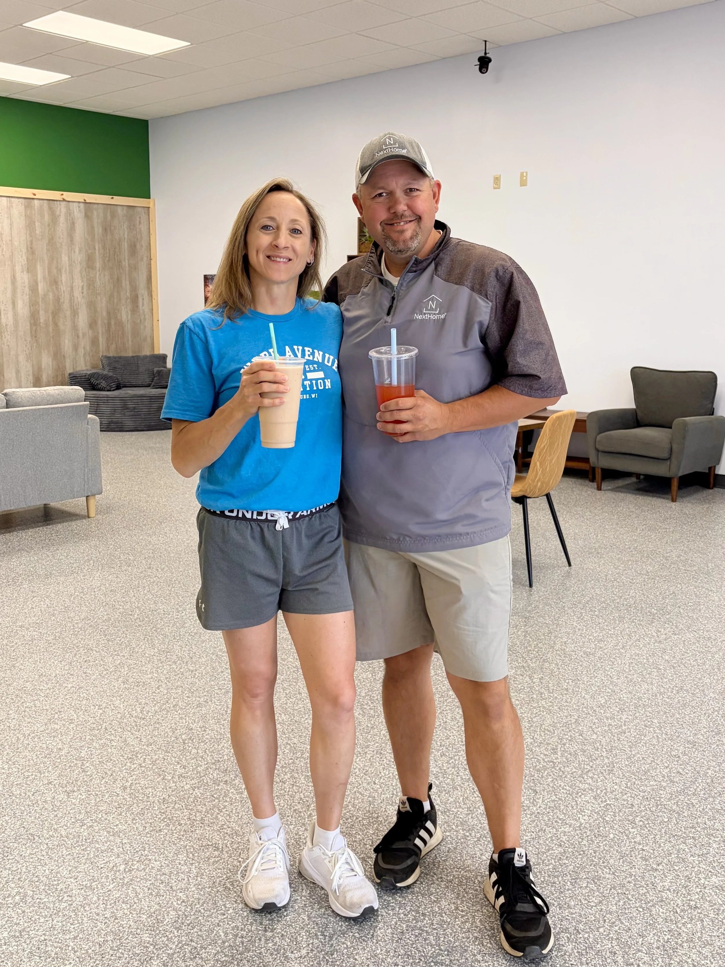 A woman and a man standing together indoors, holding drinks with straws. The woman wears a blue t-shirt and gray shorts, and the man wears a gray jacket, beige shorts, and a gray cap. They are smiling, with a seating area and wall decor in the background.