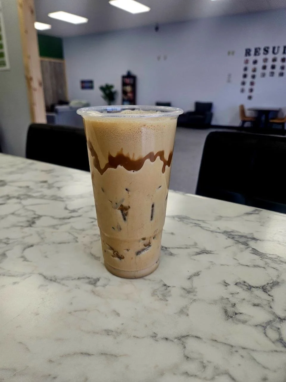 Iced coffee in a clear plastic cup with chocolate syrup drizzle, placed on a marble table in a cafe or lounge area.
