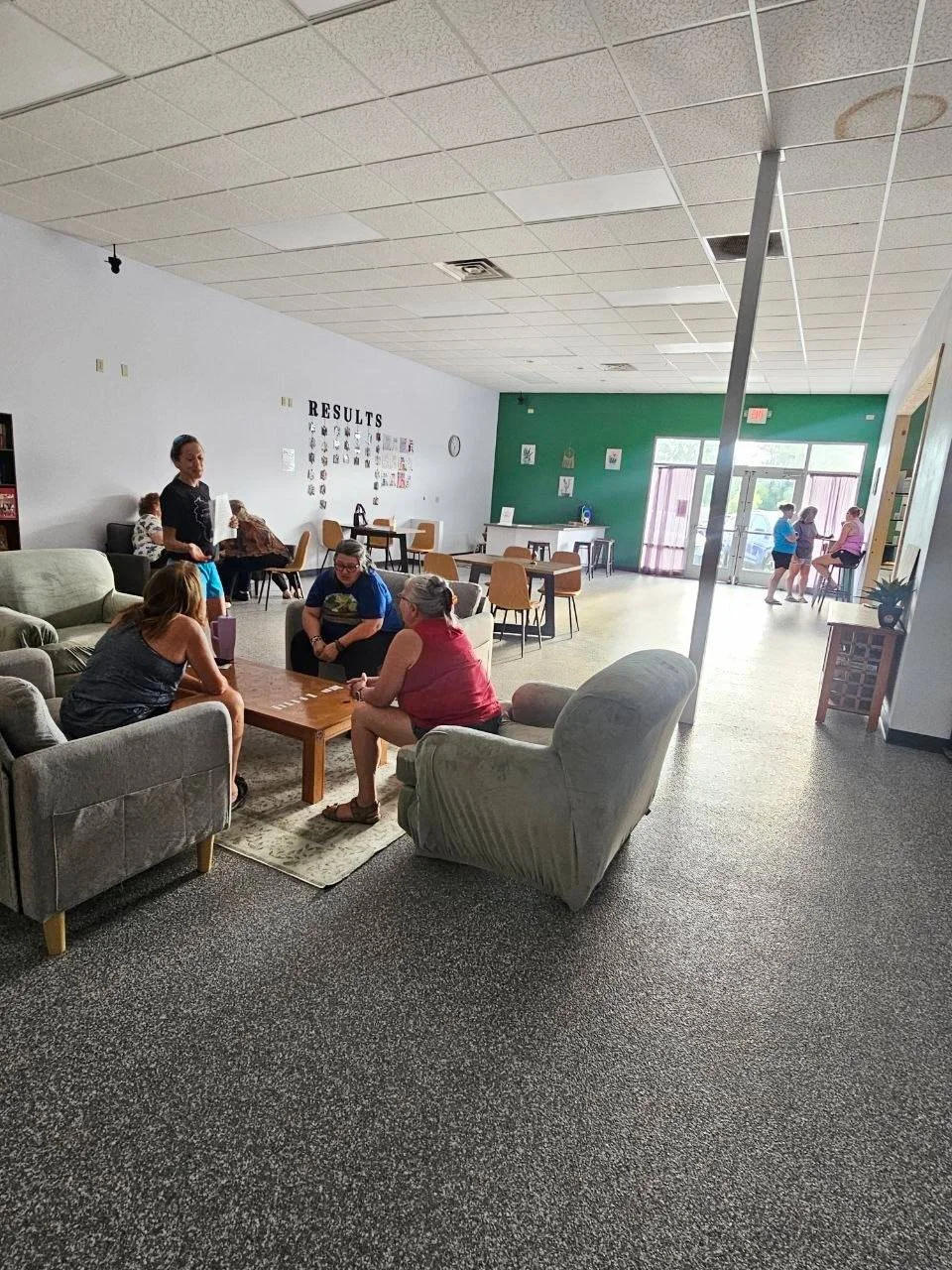 A lounge area with three women sitting on sofas and talking, a woman standing with a paper and phone, several tables and chairs, a wall with the word 'RESULTS', and a group of people near the entrance door.