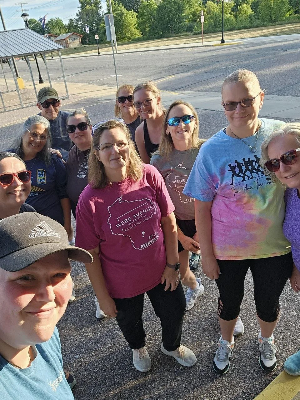 Group of women wearing athletic shirts and sunglasses, taking a selfie outdoors in a parking lot with trees and street lamps in the background.
