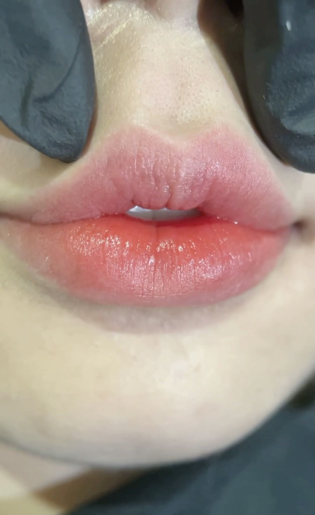 Close-up of a person's lips, showing pink lipstick and slightly parted lips. The person's nose and portions of their face are visible, with someone wearing black gloves holding the person's nose.