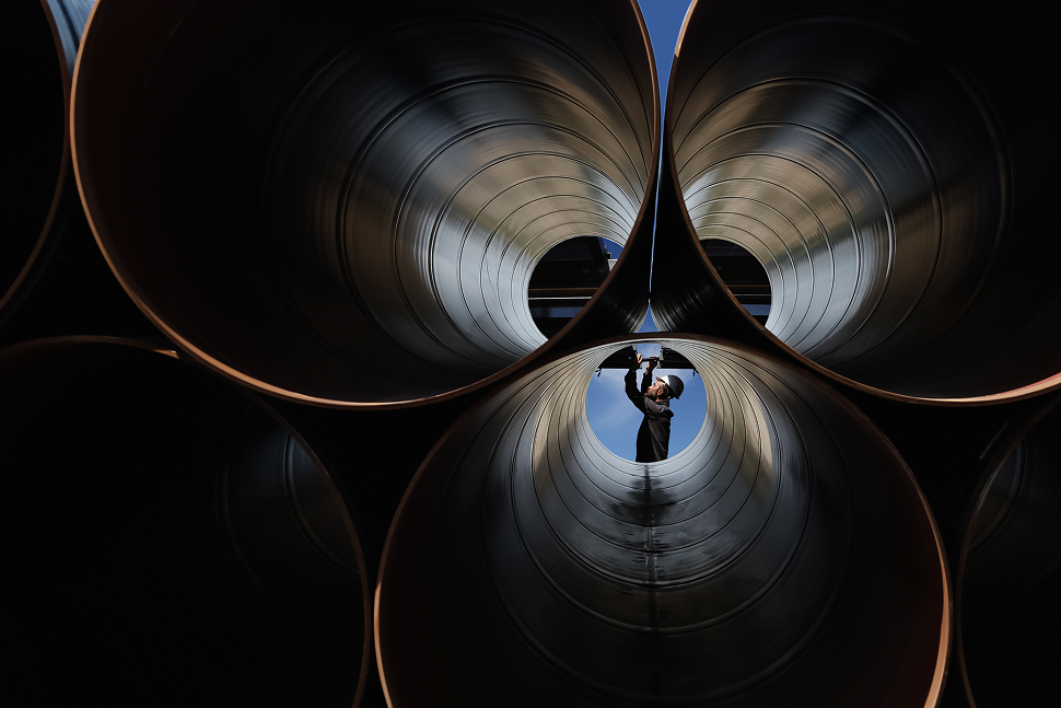 A person standing inside a large metallic pipe, reflected in the pipe's shiny interior surface, with a blue sky in the background.