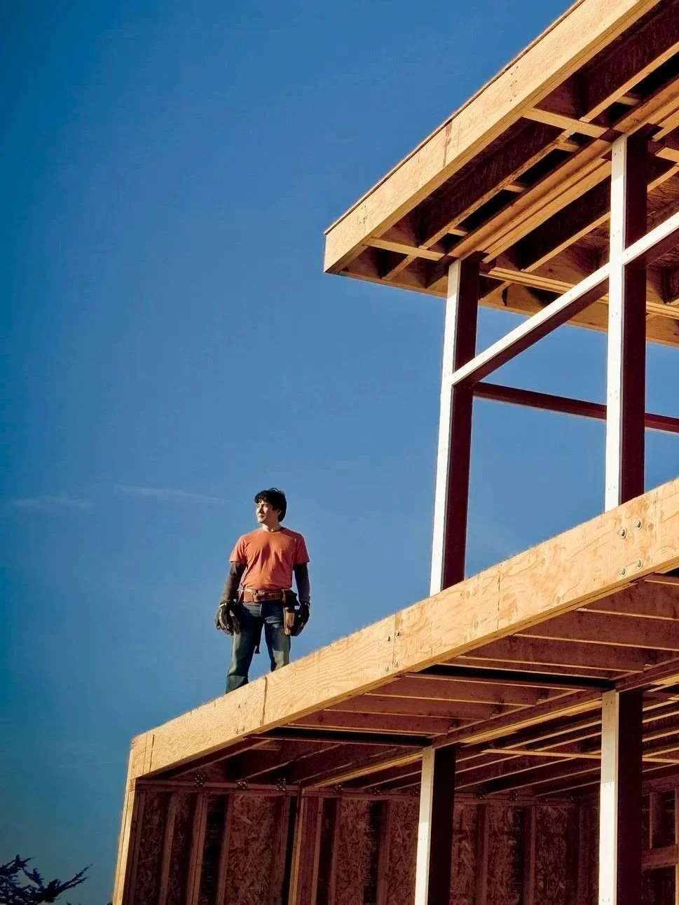 A construction worker standing on the edge of a wooden building under construction, with a clear blue sky in the background.