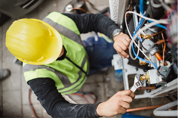 A technician inspecting or repairing electrical components and wiring on an industrial machine, wearing a yellow safety helmet and a high-visibility vest.