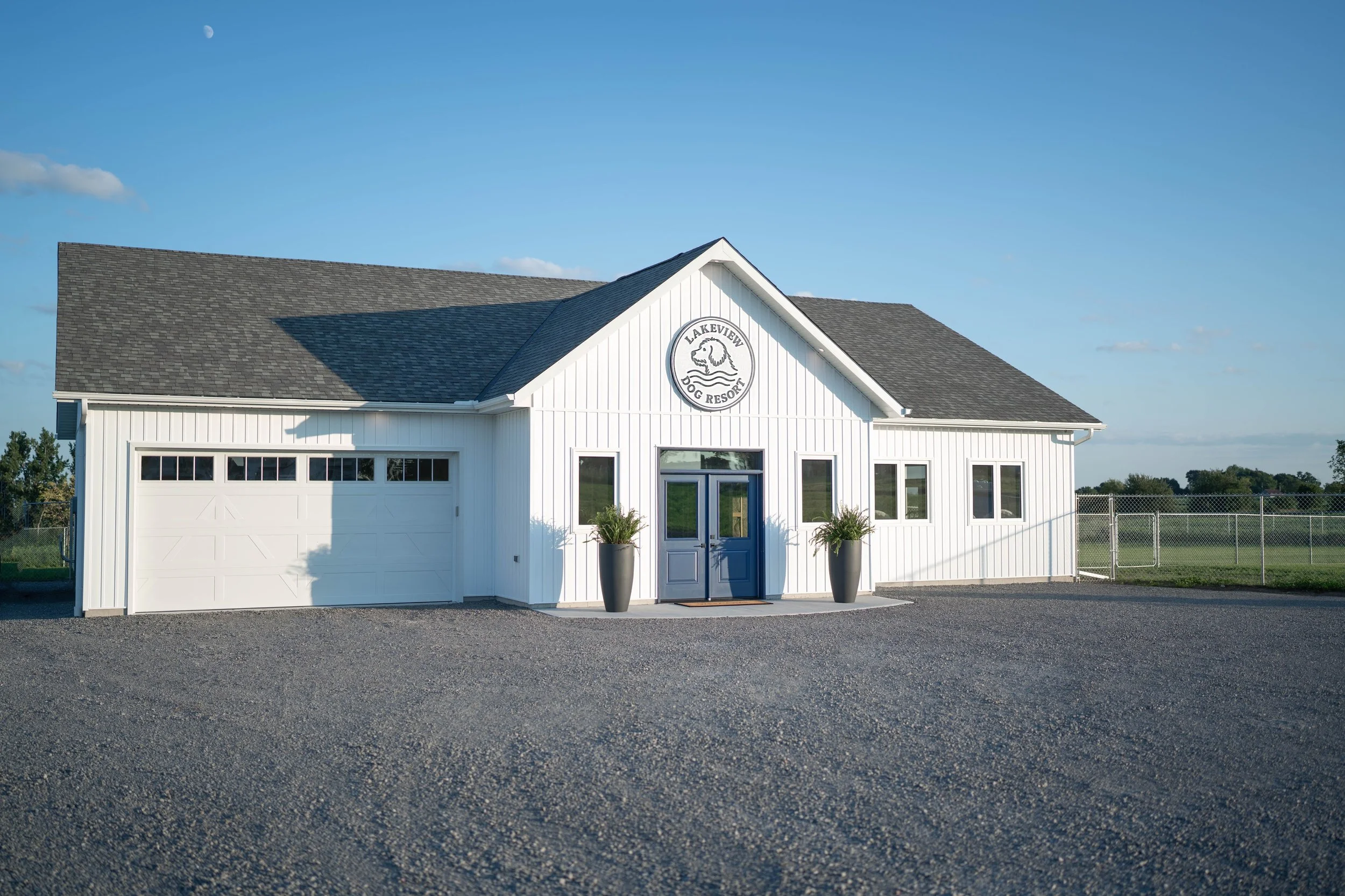White building with a sign reading 'Lakeview Dog Resort', featuring a logo of a dog's face, with two tall planters with greenery outside and a chain-link fence nearby.