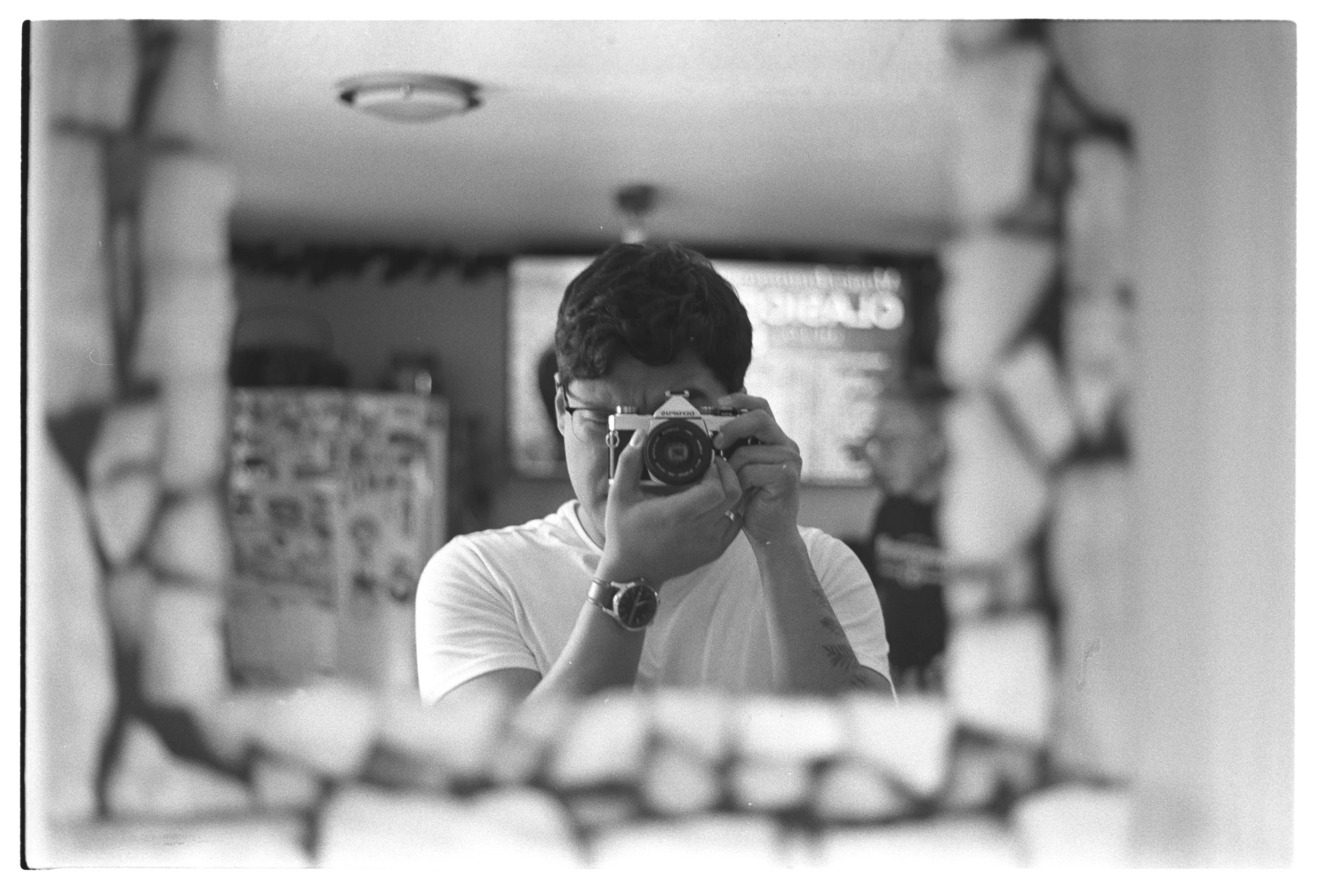 Black and white photo of a young person taking a selfie with a camera in a mirror, with signs and people in the background.