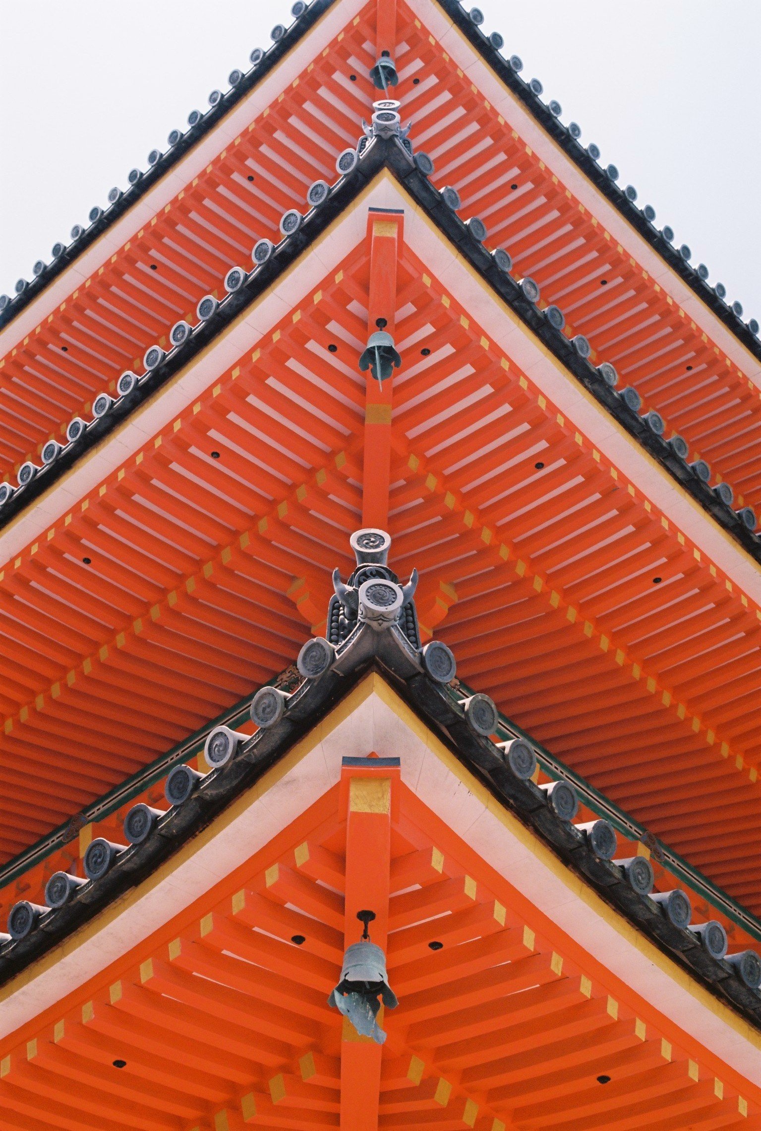 A close-up view of the corner of a traditional Japanese temple roof, featuring orange wooden beams, black tiles, decorative end caps, and hanging bells.