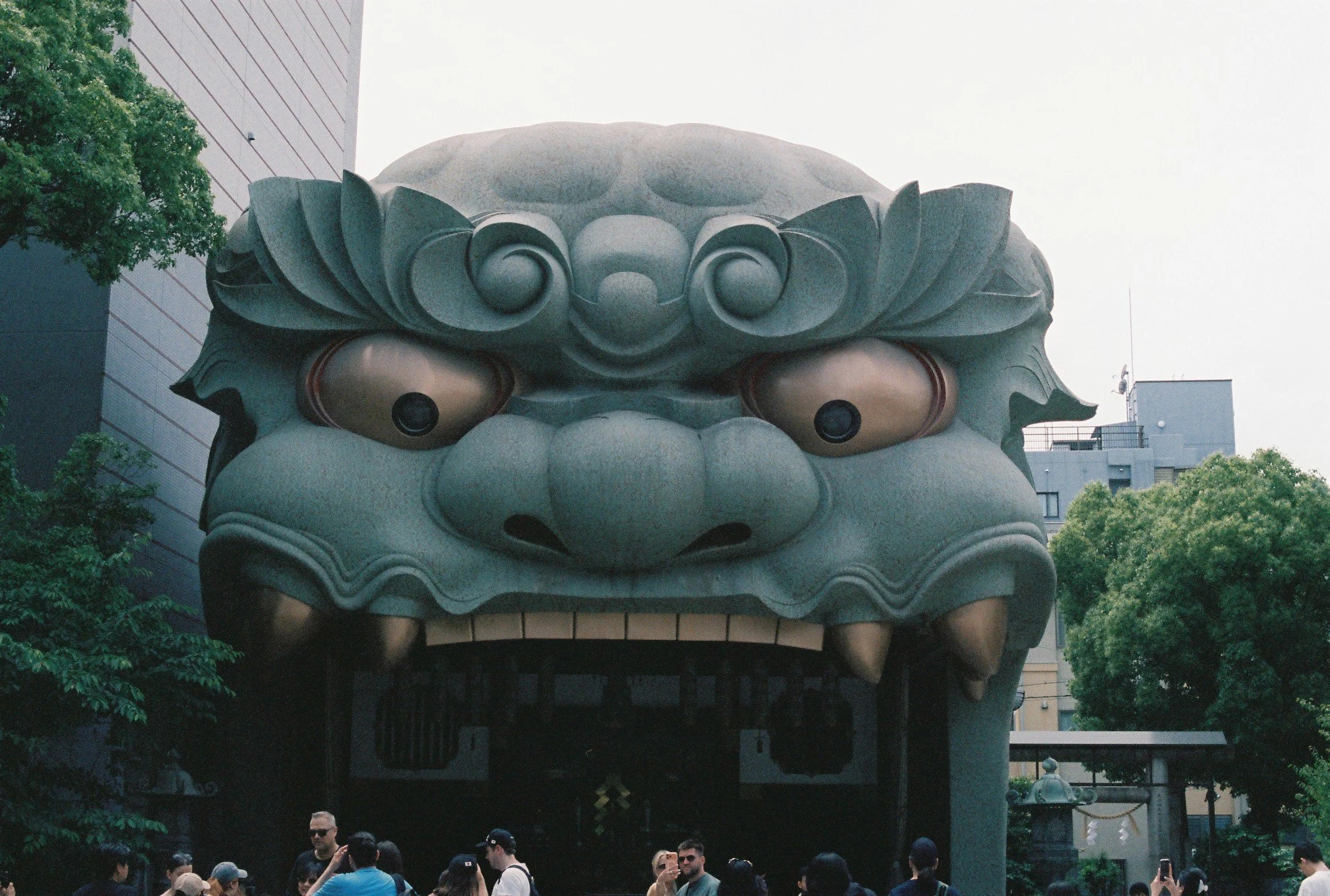 Photo of a large traditional Japanese guardian lion-dog statue, known as 'Komainu,' with a group of people gathered underneath and around it, set outdoors with trees and modern buildings in the background.