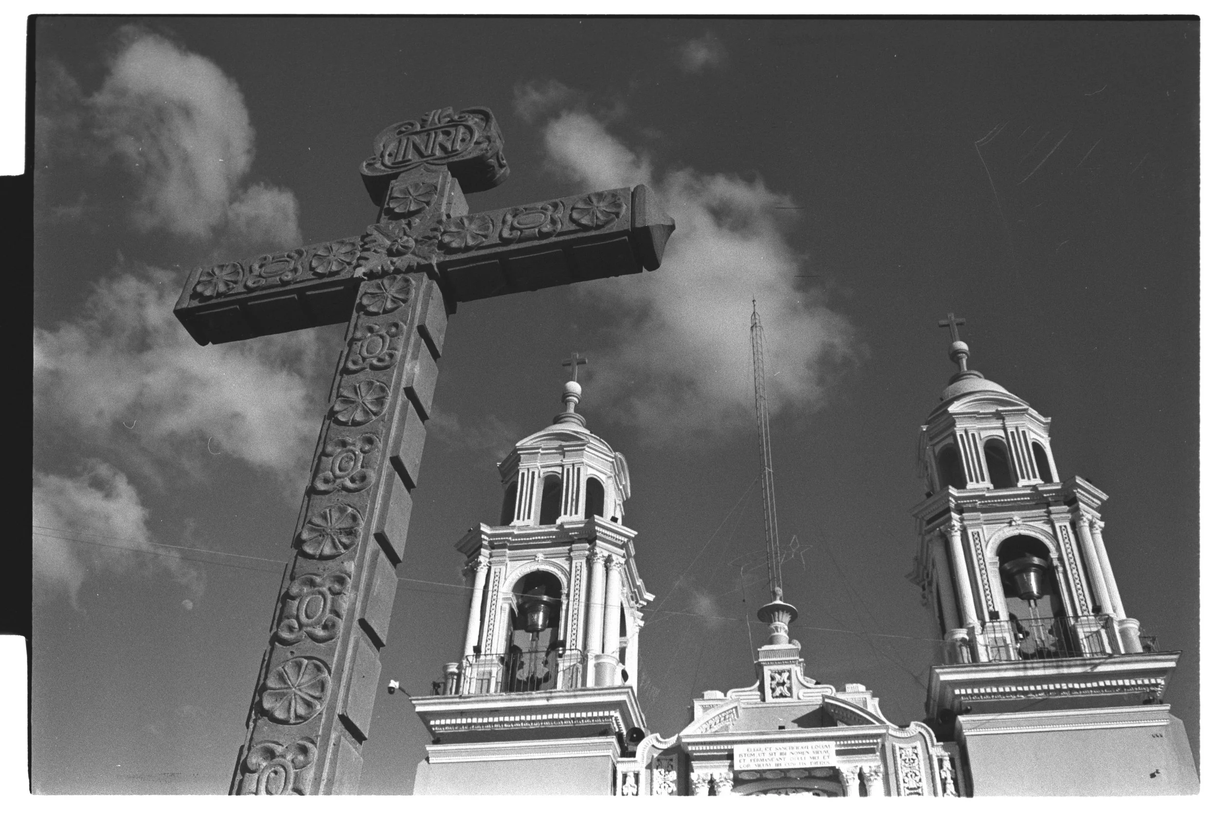 A black and white photo of a decorative cross with floral patterns in the foreground, and a church with two steeples topped with crosses in the background under a cloudy sky.