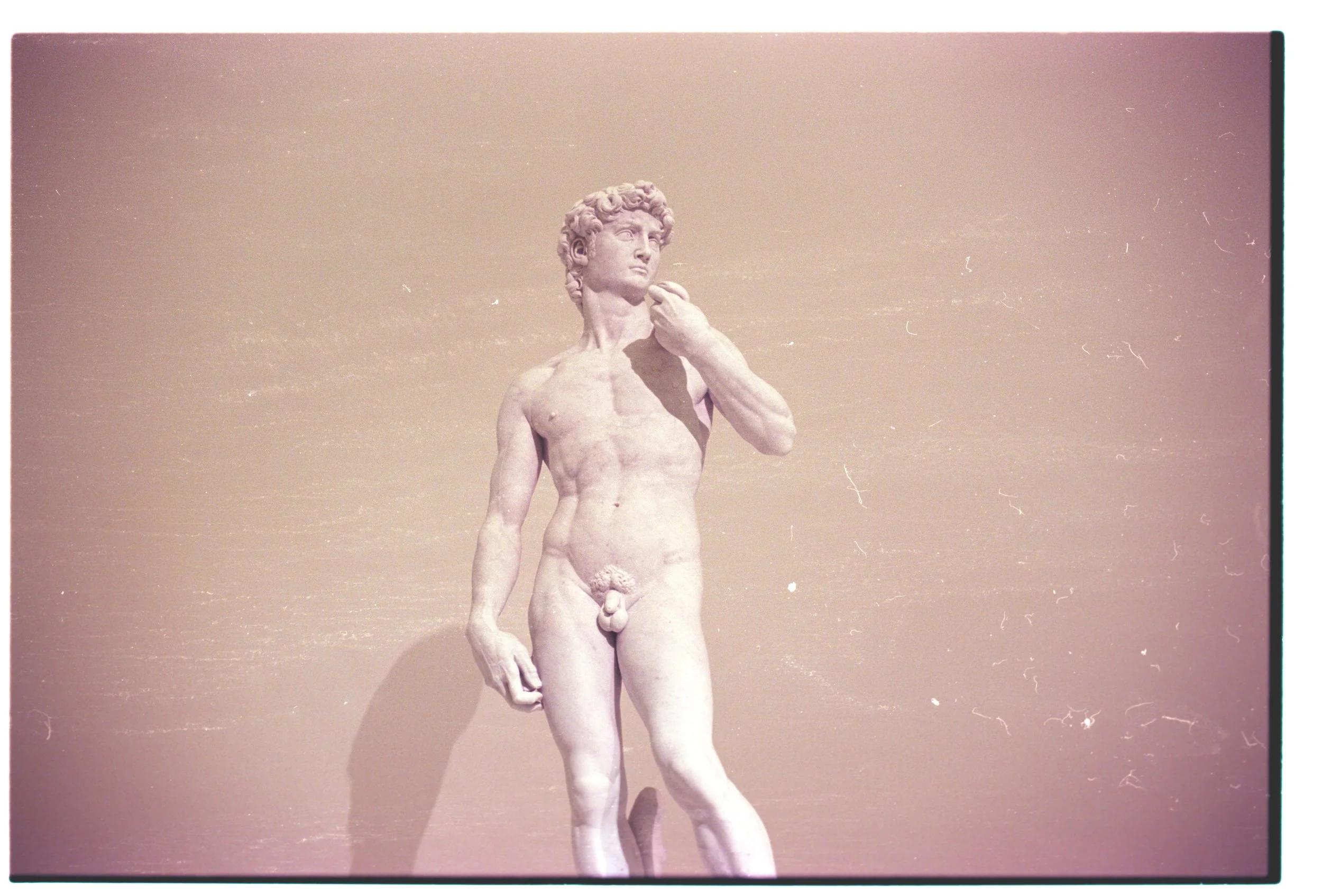 Statue of a nude male figure with curly hair, standing with one hand raised to his chin against a plain background.
