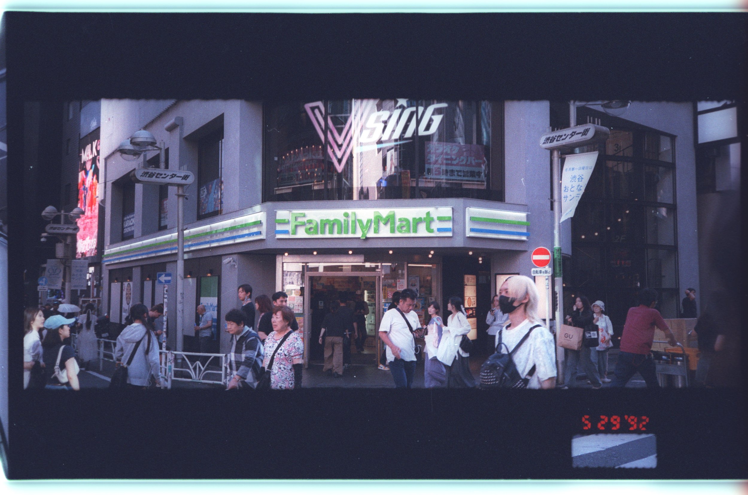 Street scene in Japan with people walking, some wearing face masks. A building with a green and white FamilyMart convenience store sign is visible. Signage in Japanese and neon advertising can be seen on the building, with a crowd gathered in front.