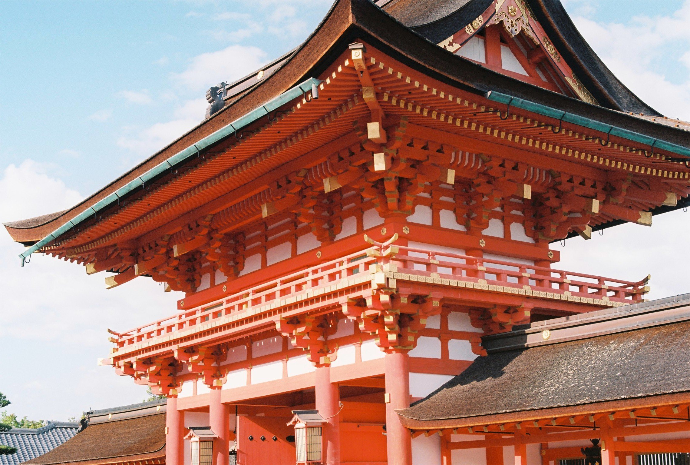 Traditional Japanese shrine building with red and white wooden structure, detailed roof, and decorative elements, set against a blue sky with clouds.