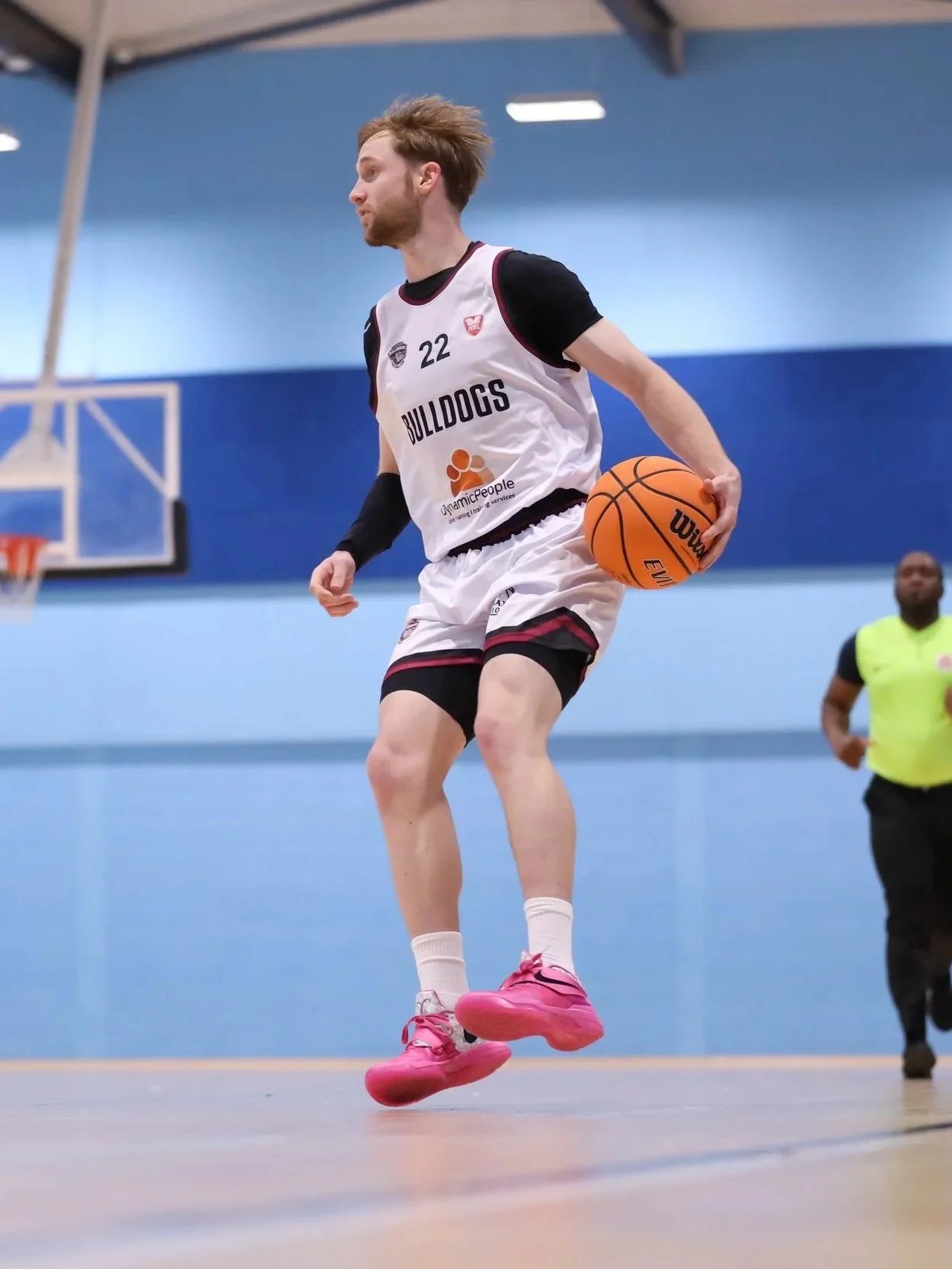 A young man in a basketball uniform with the number 22 is jumping and dribbling a basketball on an indoor court. He is wearing pink sneakers and has light brown hair. In the background, a referee or coach in a yellow shirt is standing, and a basketball hoop is visible.