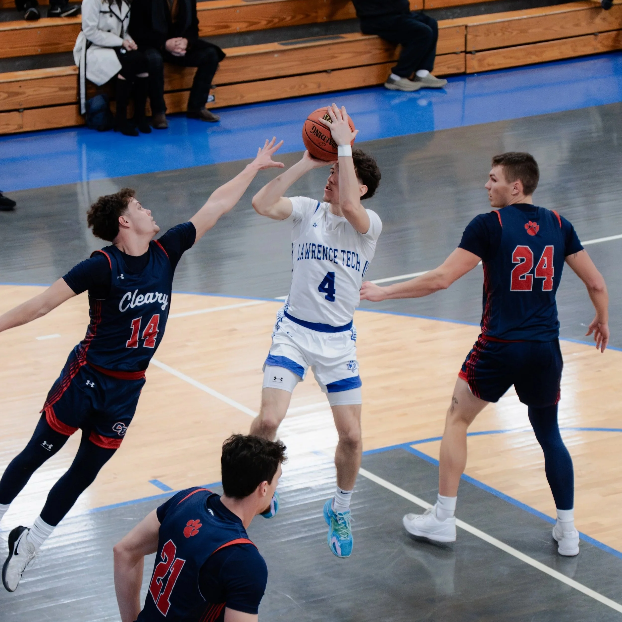 Two high school basketball players, one in a white team uniform from Lawrence Tech and the other in a blue uniform, are competing for possession of the basketball during a game, with one player reaching up to grab it.