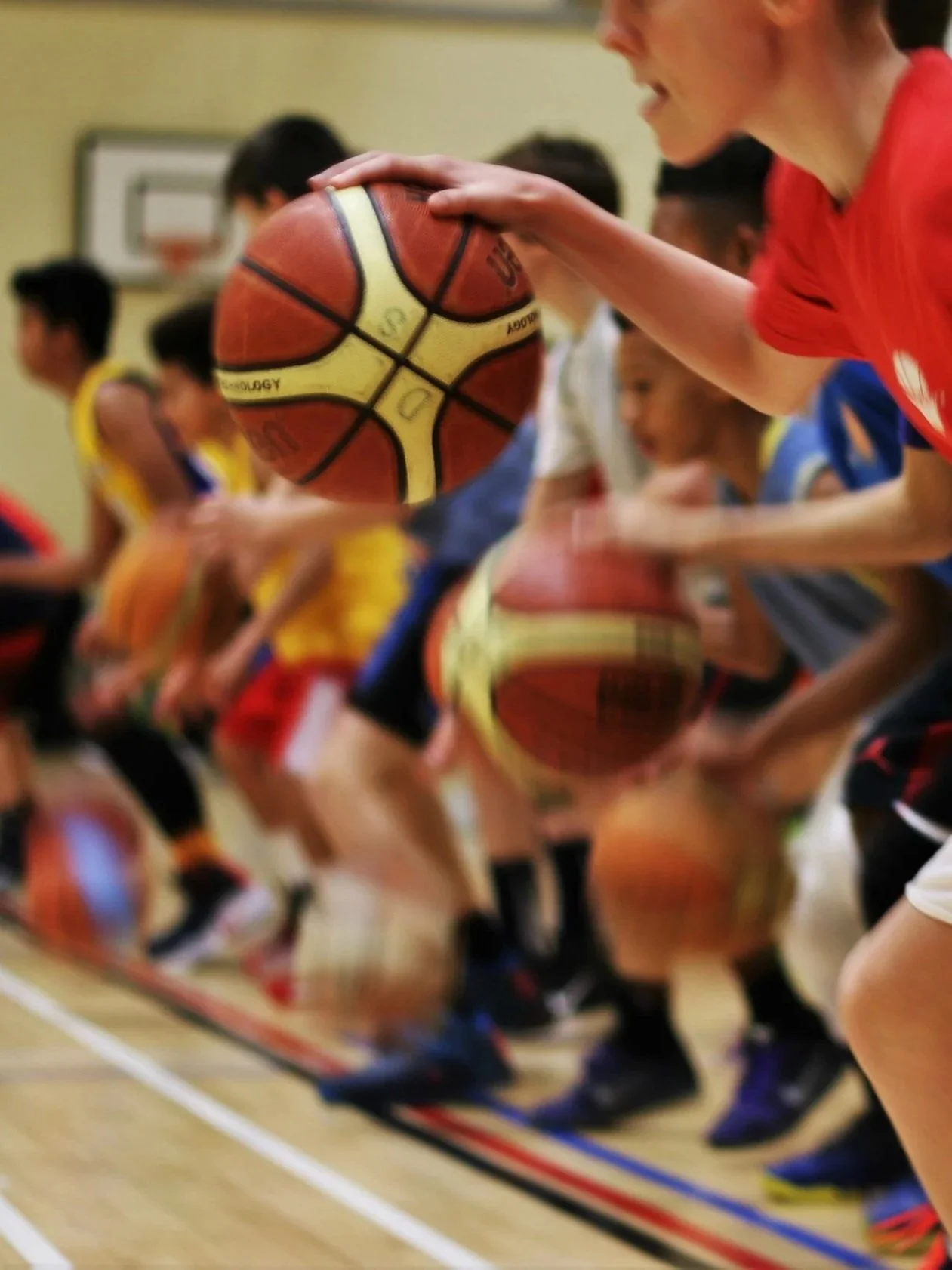 A group of children participating in a basketball drill, each holding a basketball, in a gymnasium with a basketball hoop in the background.