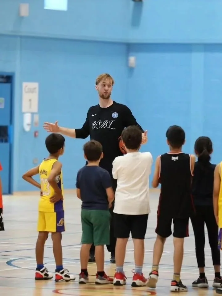 A basketball coach is speaking to a group of young children in a gymnasium, with the players wearing different team uniforms and standing in a semi-circle.
