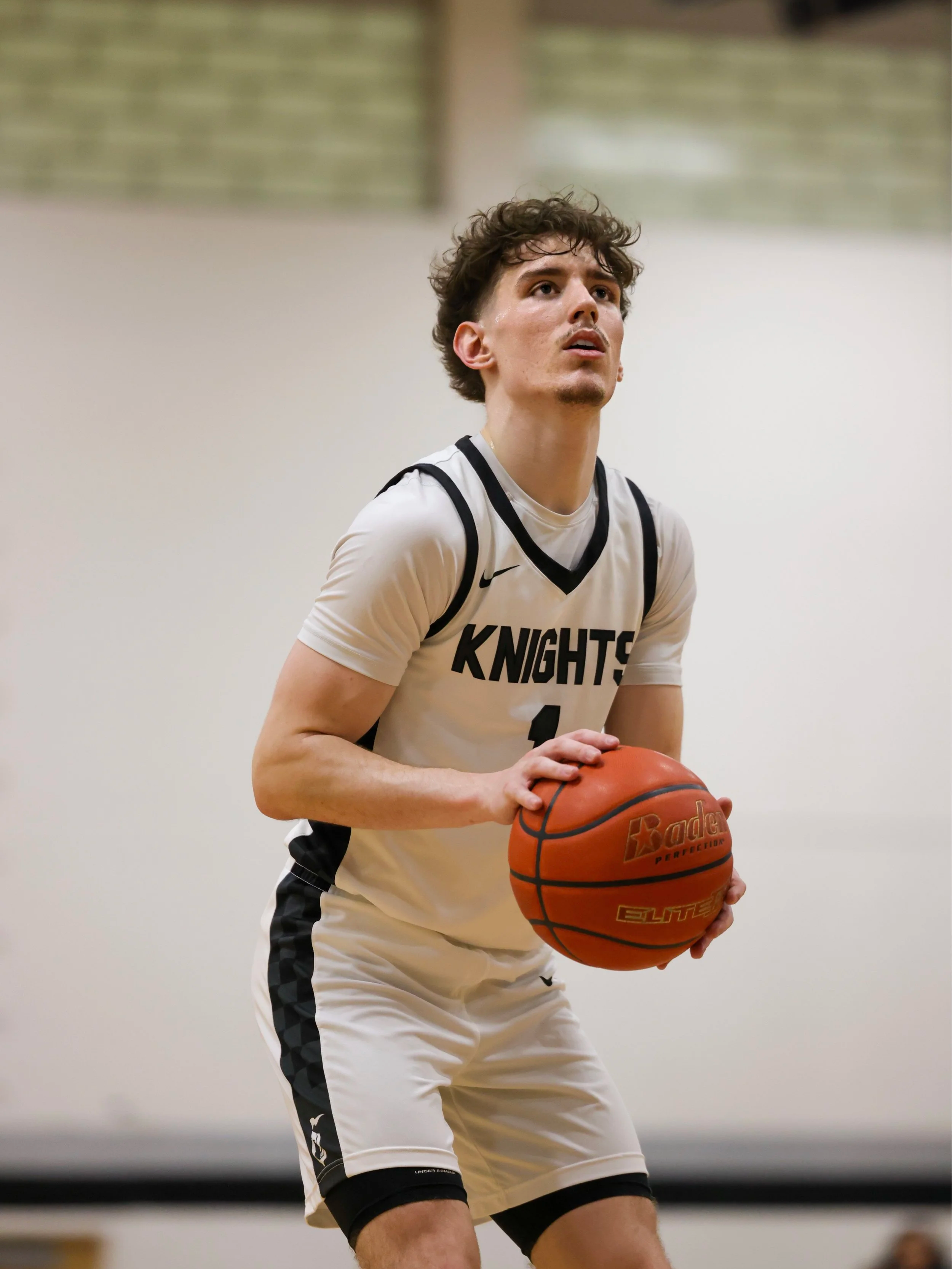 A male basketball player holding a basketball, wearing a cream-colored jersey with black accents that reads "KNIGHTS," in an indoor gym or basketball court.