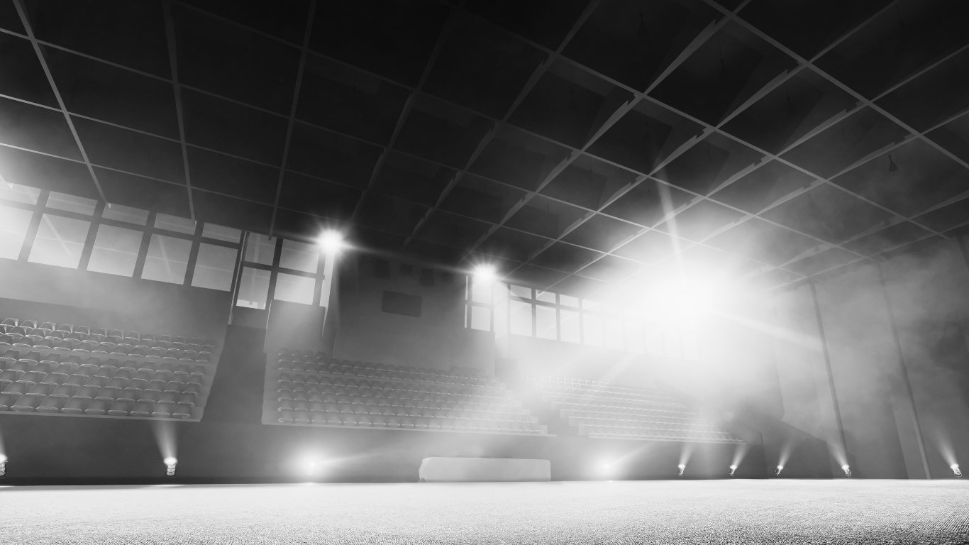 Empty indoor sports arena or theater with bright lights, rows of seating, and a stage or floor area, illuminated with fog or smoke for a performance or event.