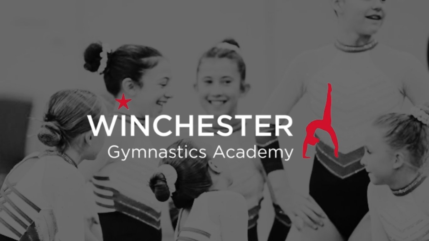 Black and white photo of young female gymnasts in uniforms, gathered and smiling, with the logo of Winchester Gymnastics Academy overlayed, featuring a red star and a stylized gymnast.
