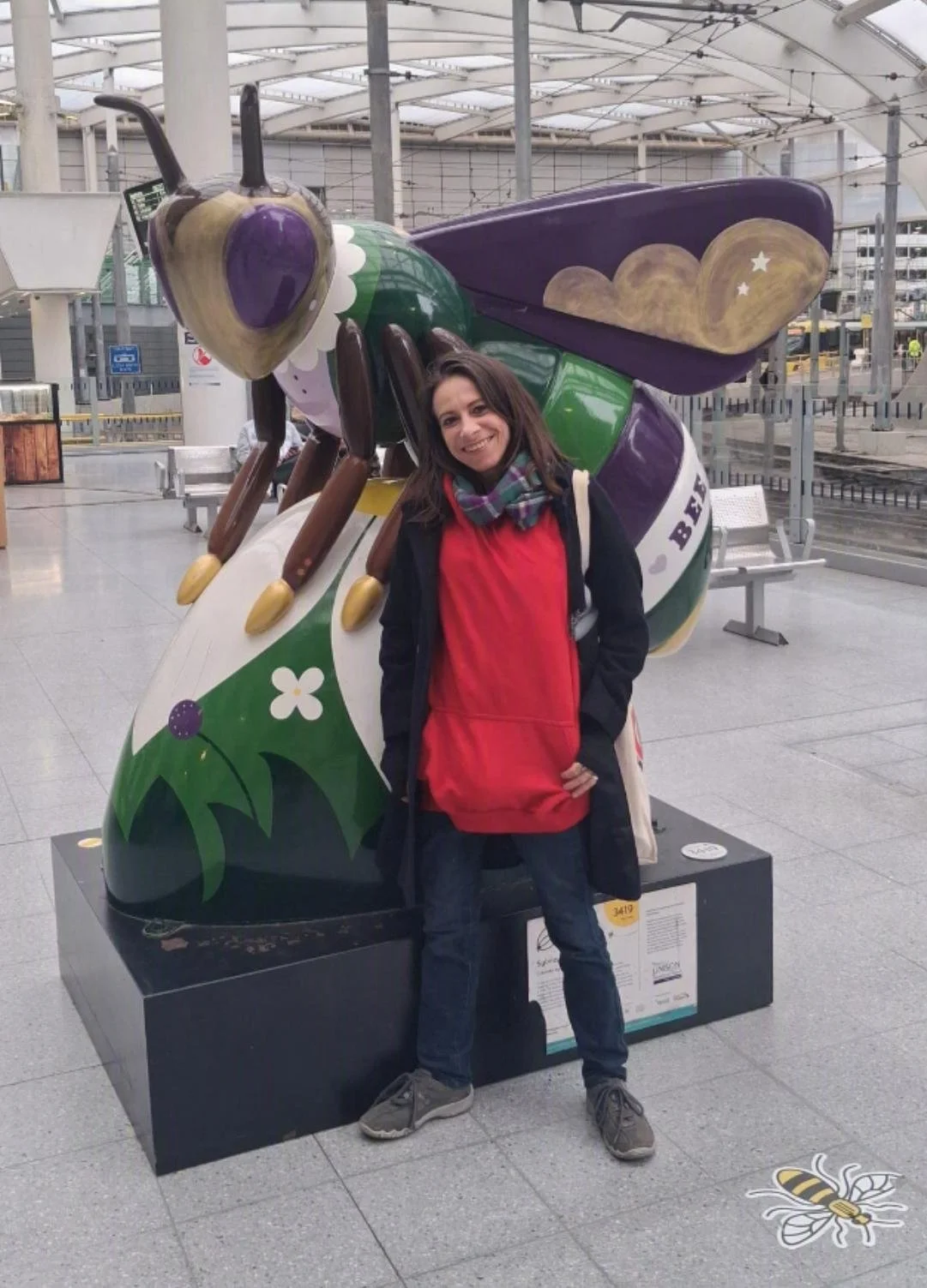A woman standing in front of a colorful bee sculpture inside a modern building with glass and steel architecture.